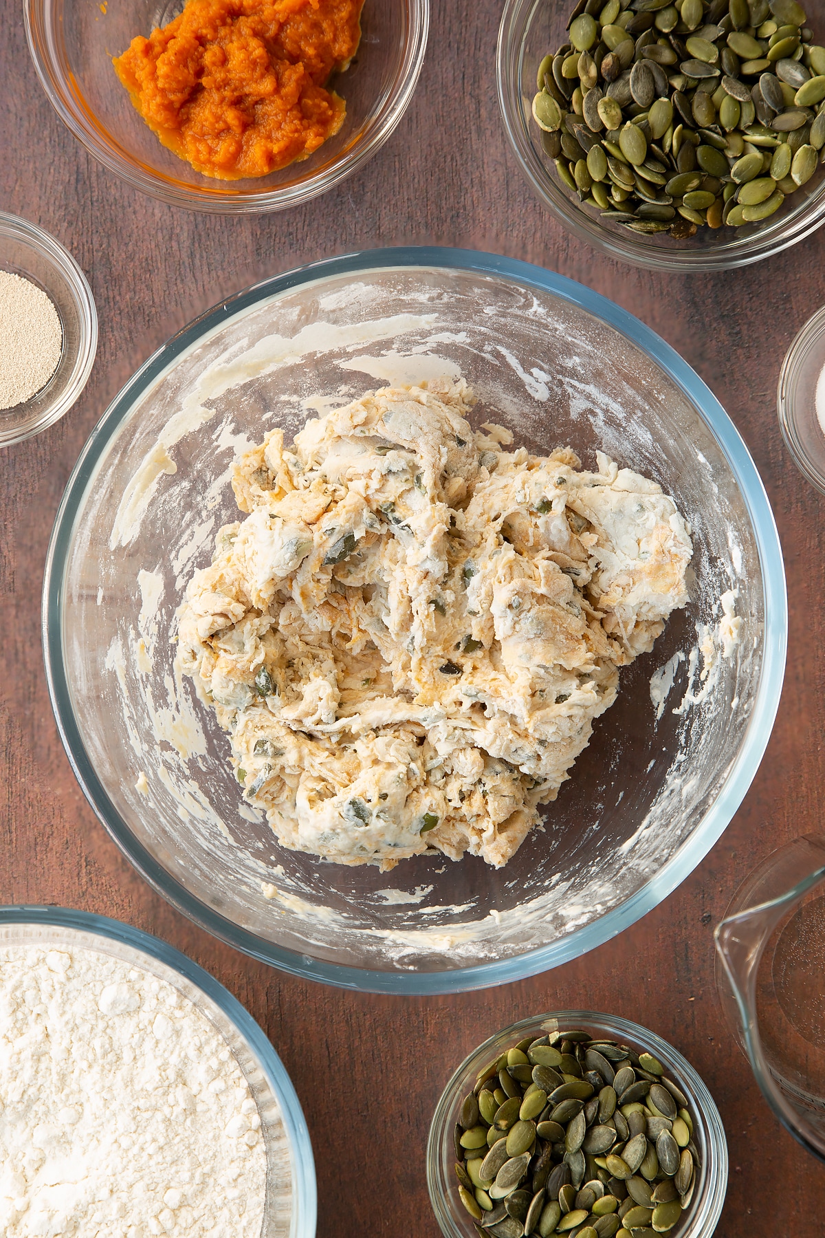 Rough pumpkin seed bread dough in a glass mixing bowl. Ingredients to make a pumpkin seed bread recipe surround the bowl.