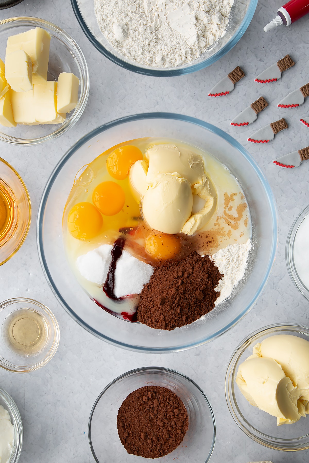 Margarine, milk, sugar, flour, cocoa, eggs and food colouring in a glass bowl. Ingredients to make red velvet Halloween cupcakes surround the bowl.