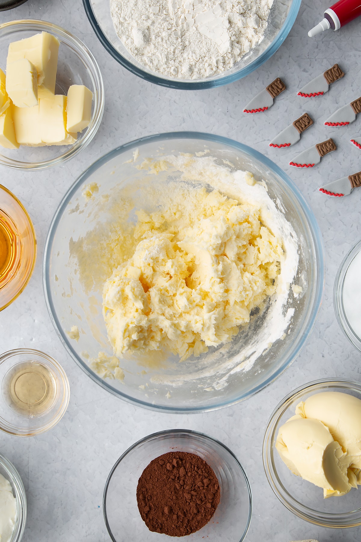 Butter, icing sugar and vanilla extract roughly mixed together in a glass bowl. Ingredients to make red velvet Halloween cupcakes surround the bowl.