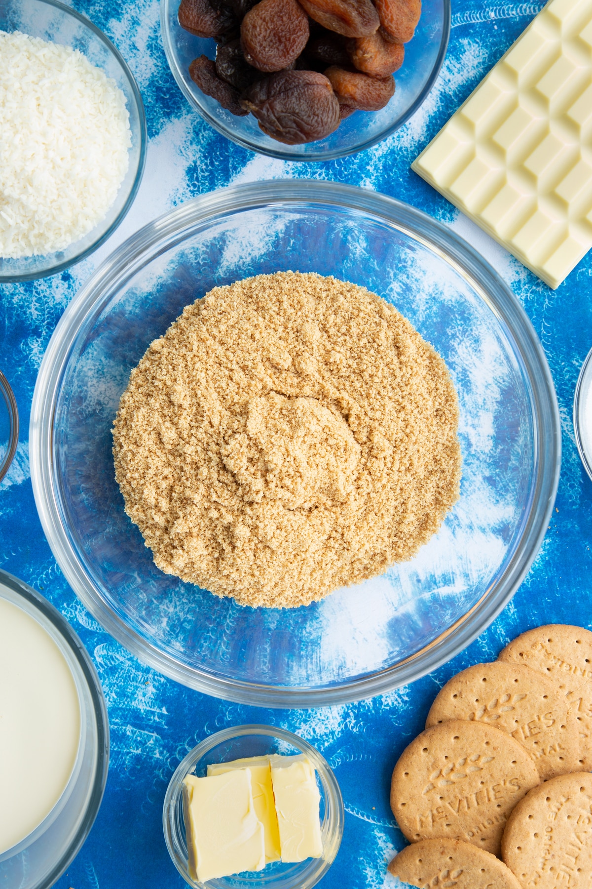 Crushed biscuits in a bowl. Ingredients to make an apricot cheesecake surround the bowl.