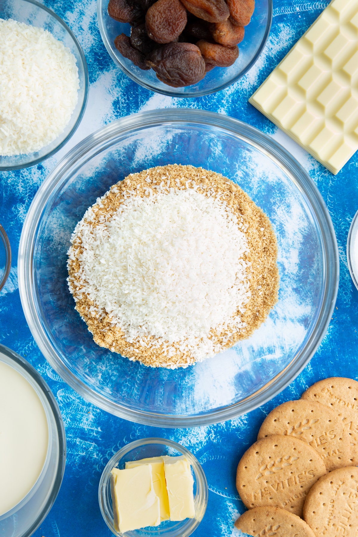 Crushed biscuits and desiccated coconut in a bowl. Ingredients to make an apricot cheesecake surround the bowl.