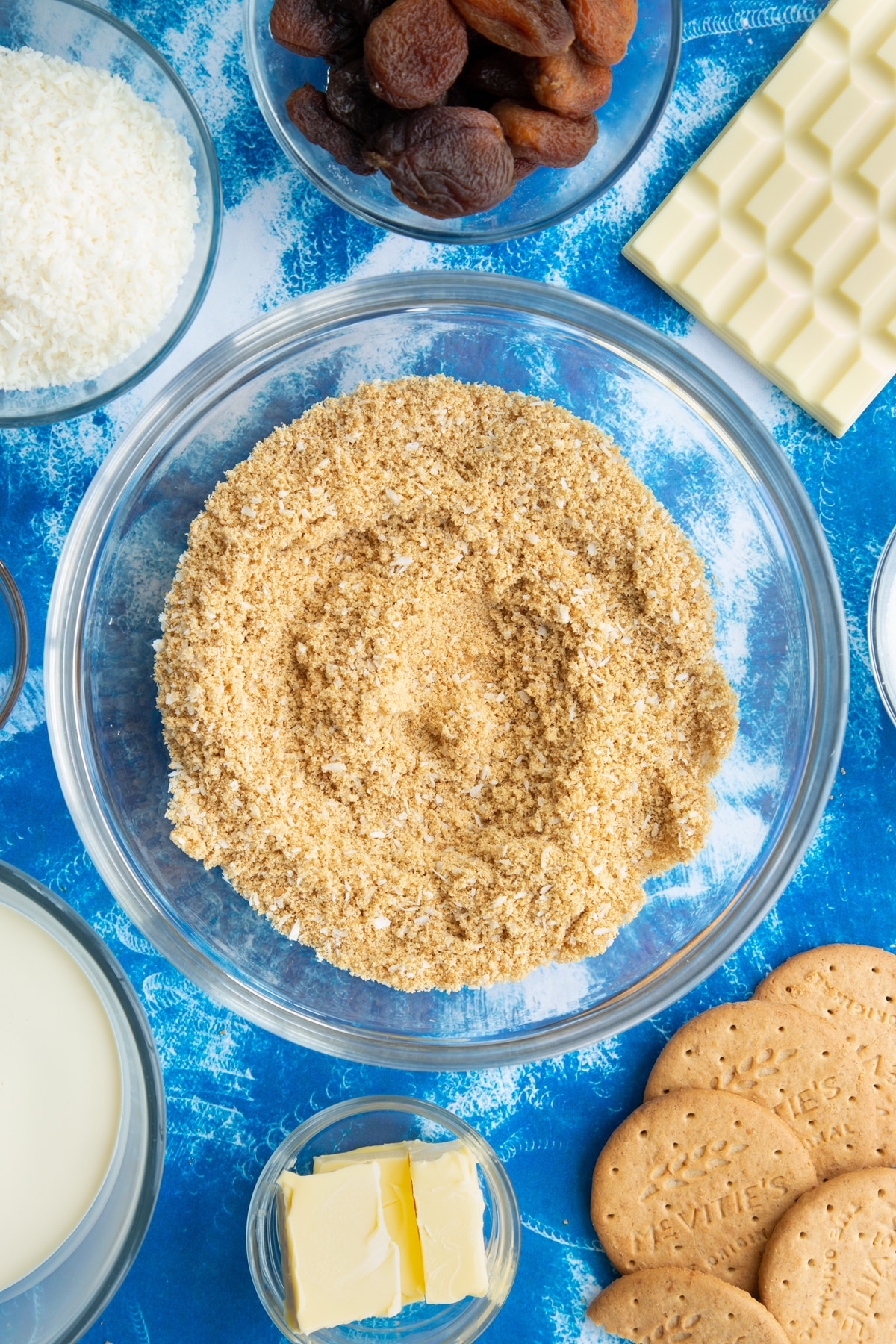 Crushed biscuits and desiccated coconut mixed together in a bowl. Ingredients to make an apricot cheesecake surround the bowl.