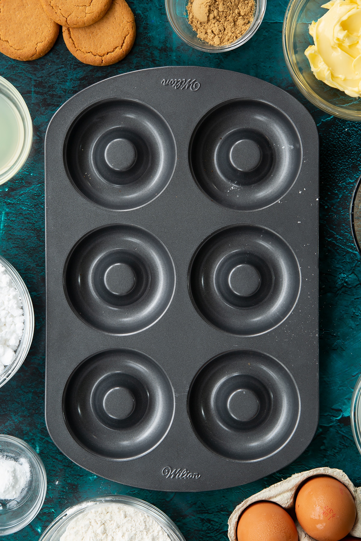 A metal donut pan. Ingredients to make gingerbread donuts surrounds the pan.