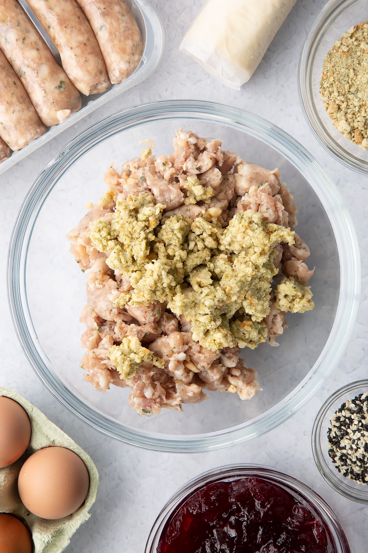 Sausage meat and stuffing in a glass mixing bowl. Ingredients to make a festive sausage roll recipe surround the bow.