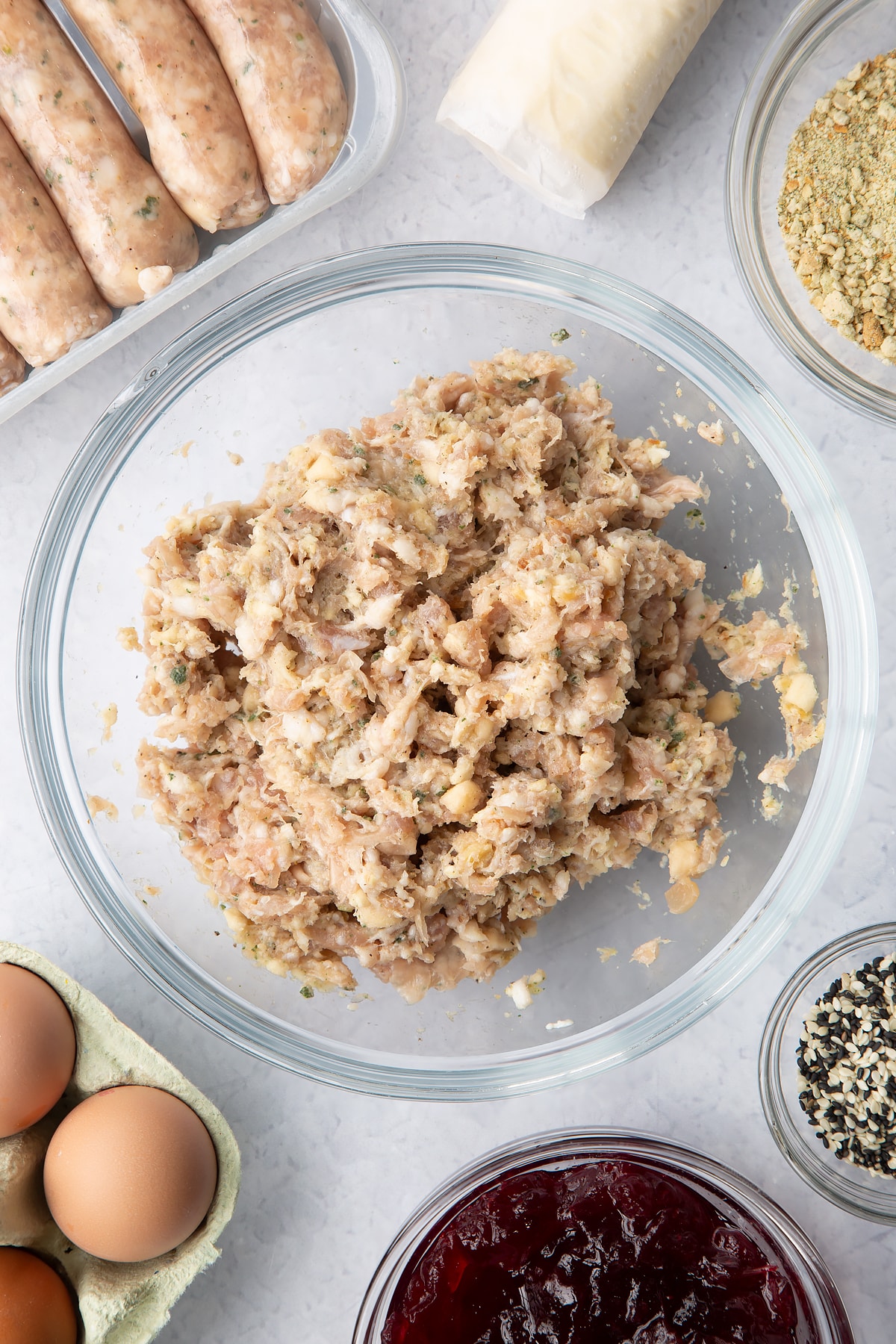 Sausage meat and stuffing mixed together in a glass mixing bowl. Ingredients to make a festive sausage roll recipe surround the bow.