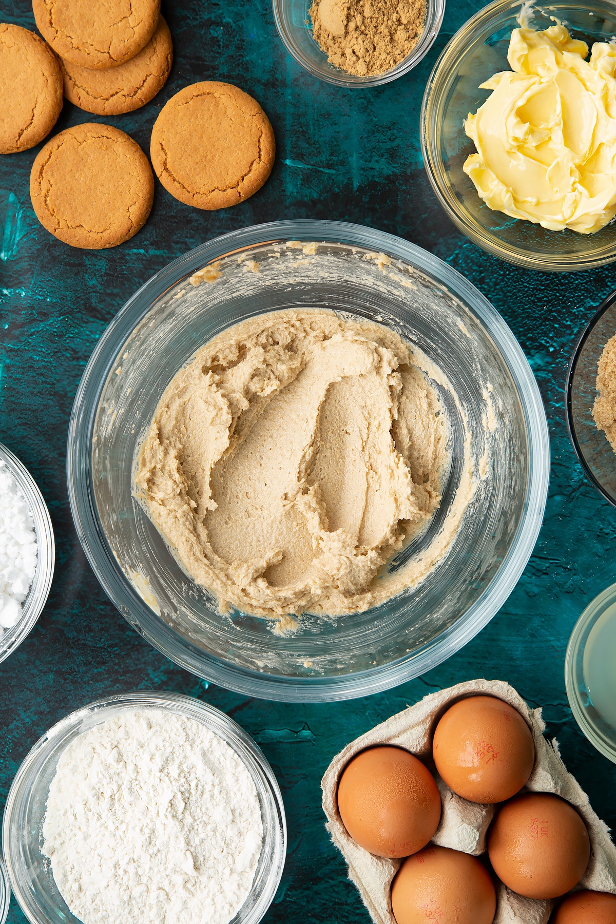 Butter and brown sugar beaten together in a glass mixing bowl. Ingredients to make gingerbread donuts surround the bowl.