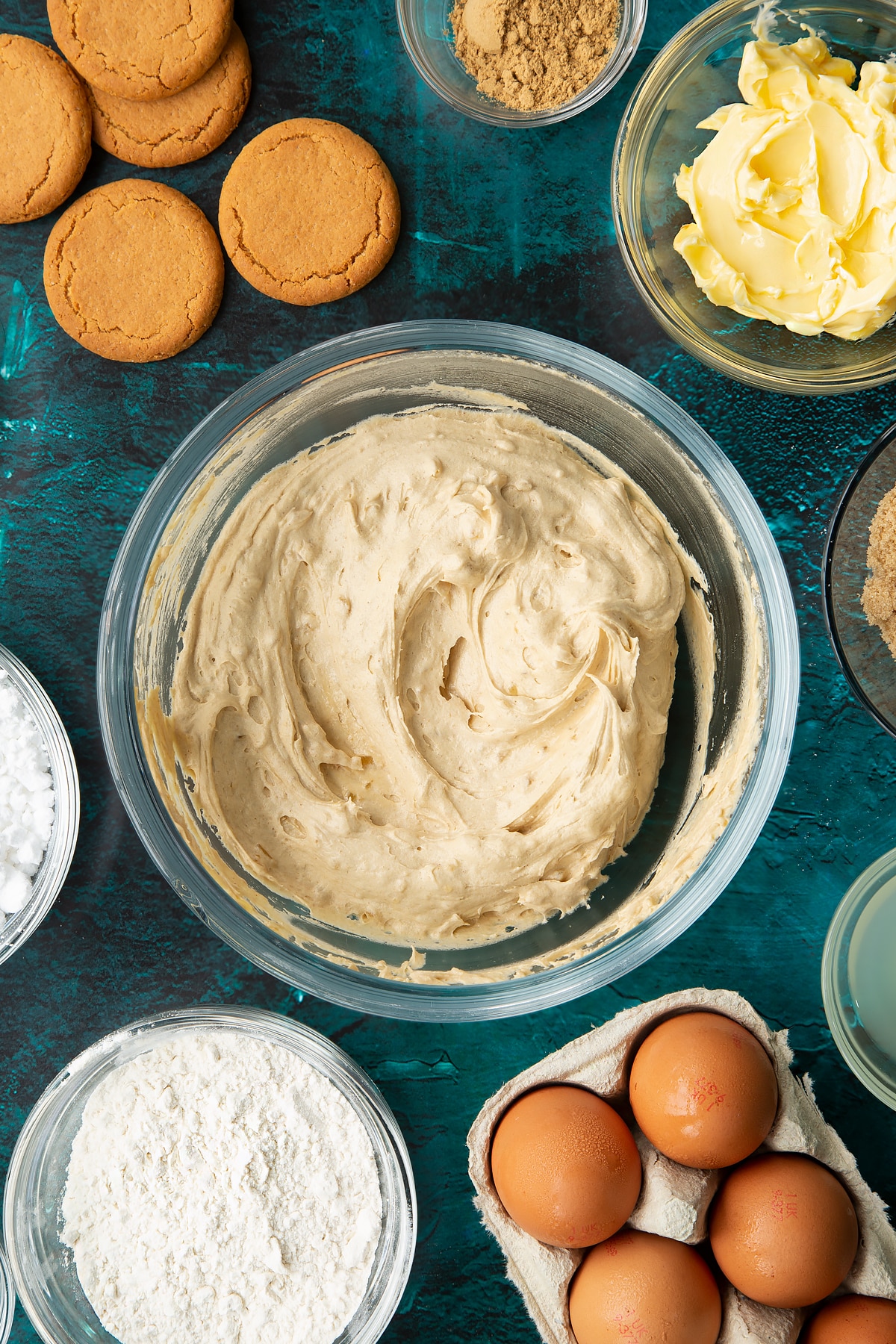 Gingerbread donut batter in a glass mixing bowl. Ingredients to make gingerbread donuts surround the bowl.