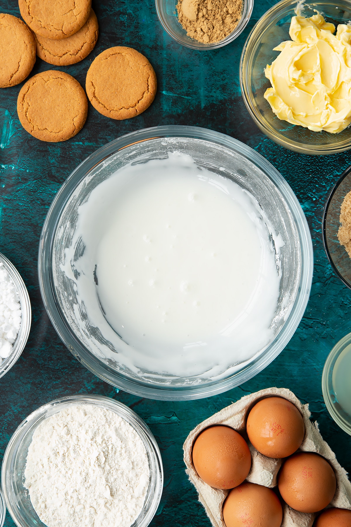 Lemon icing in a glass mixing bowl. Ingredients to make gingerbread donuts surround the bowl.