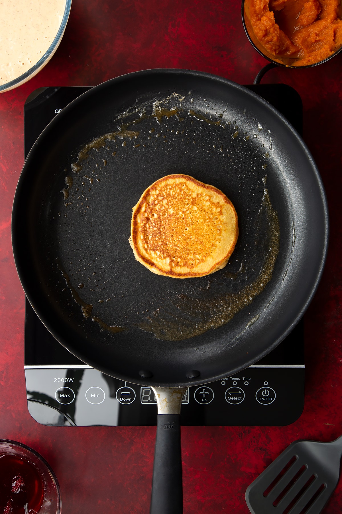 A partially cooked pumpkin pancake in a greased frying pan. Ingredients to make Halloween pancakes surround the pan.