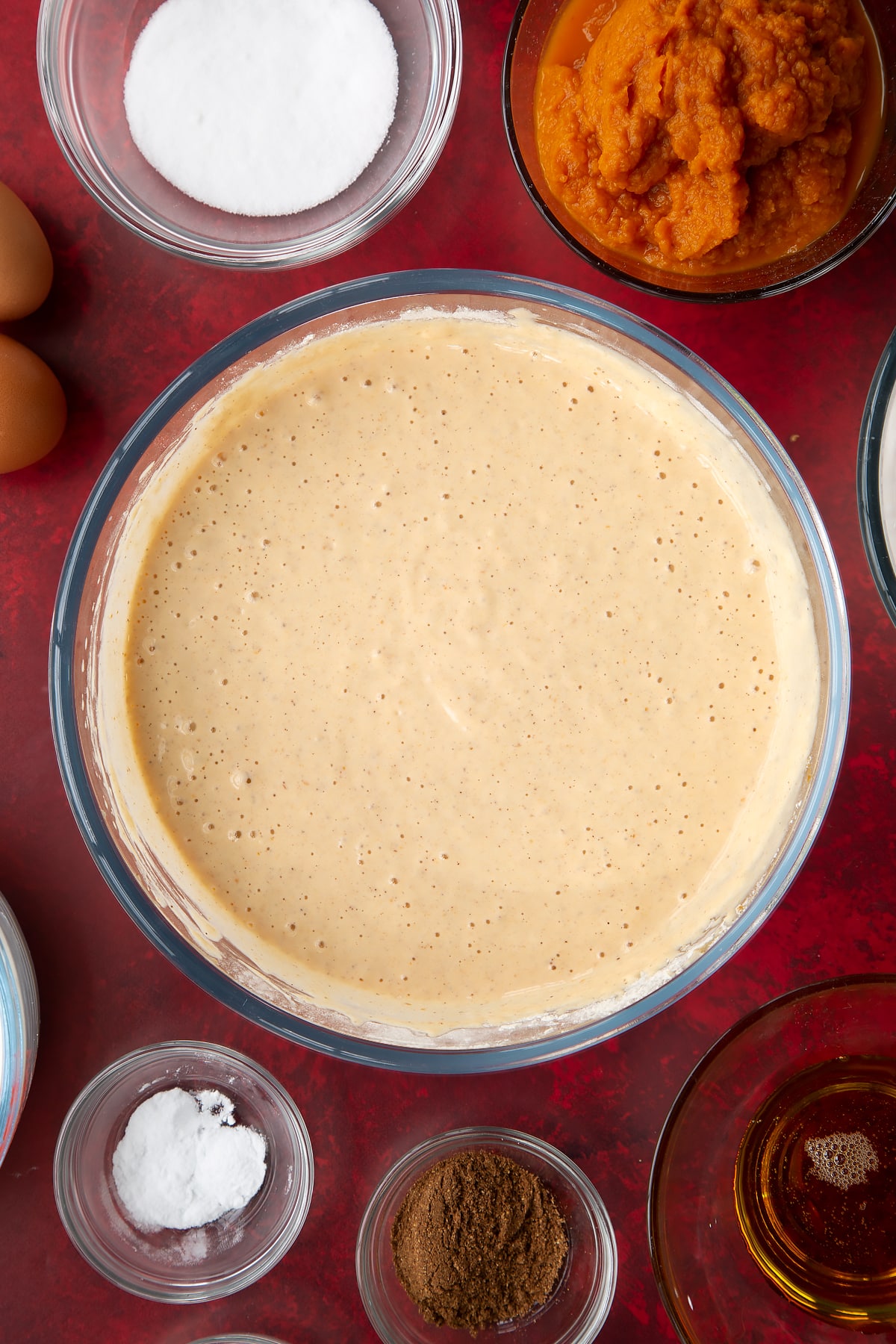 Pumpkin pancake batter in a glass mixing bowl. Ingredients to make Halloween pancakes surround the bowl.