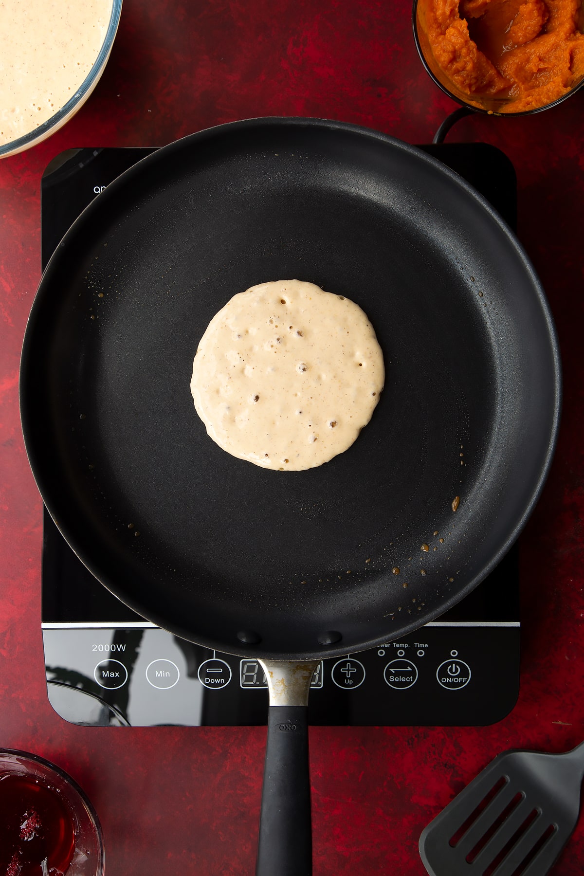 Pumpkin pancake batter bubbling in a greased frying pan. Ingredients to make Halloween pancakes surround the pan.