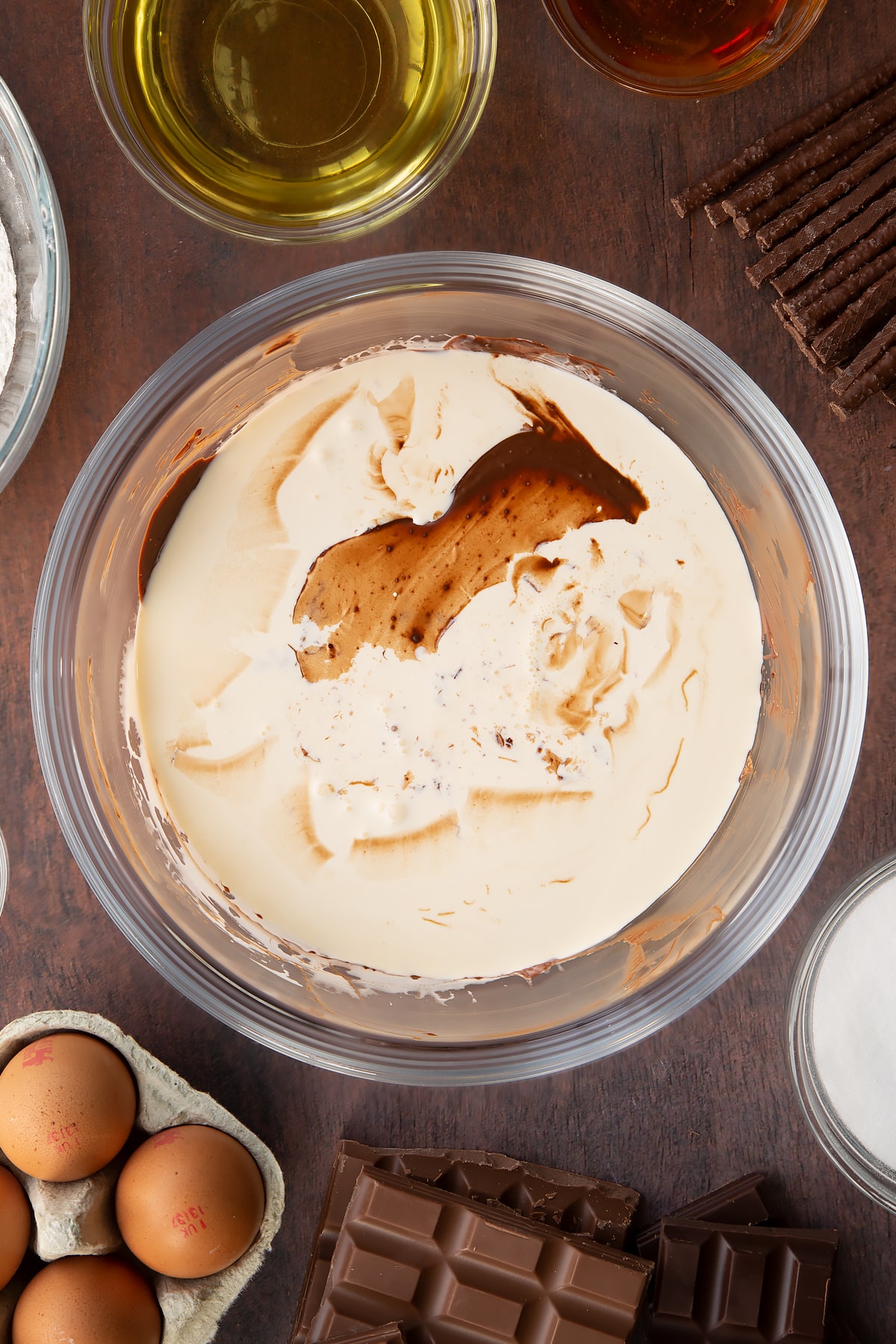 Melted dark chocolate, milk chocolate and Matchmakers in a glass mixing bowl with cream on top. Ingredients to make Matchmaker cake surround the bowl.