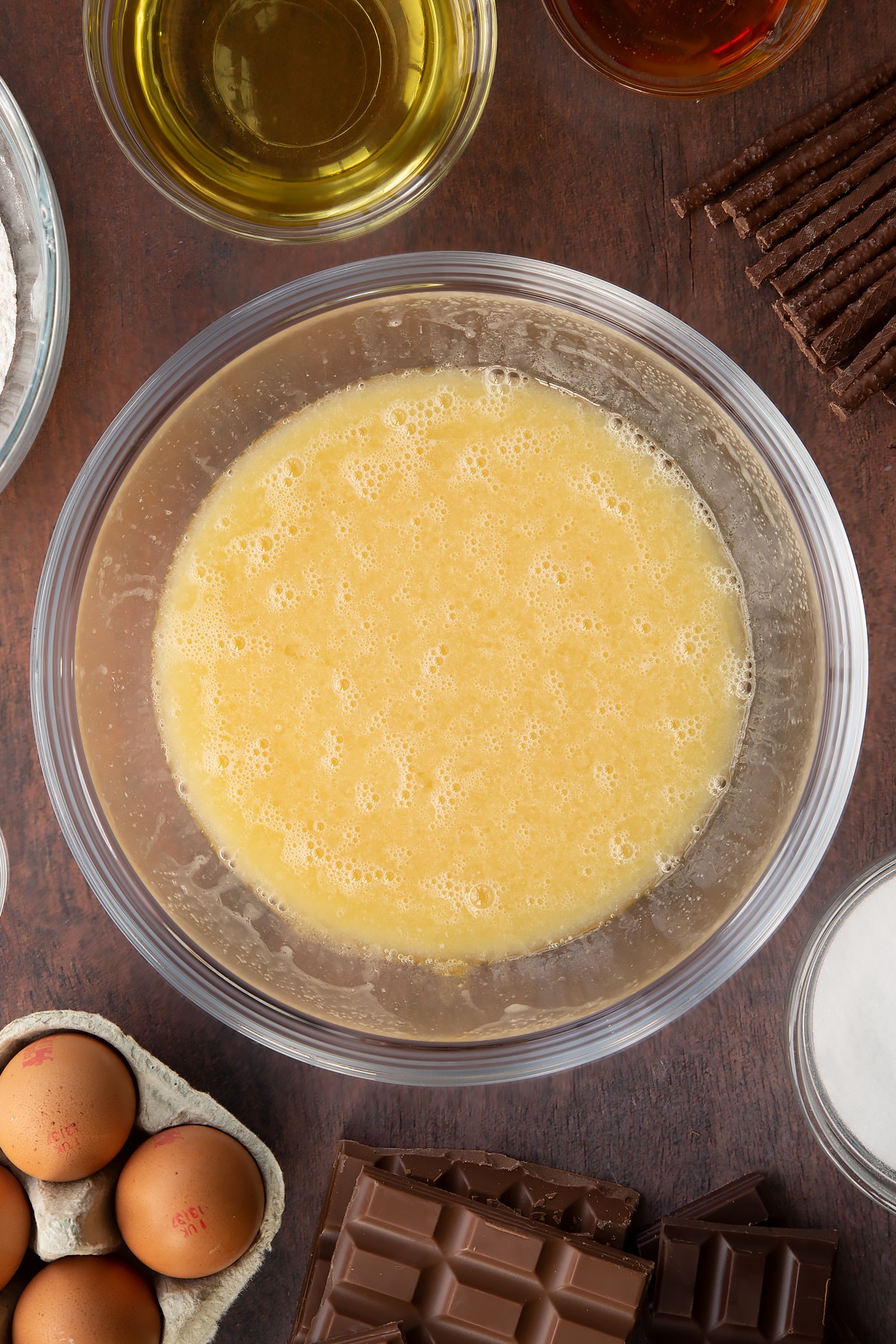 Sugar, beaten eggs, oil, milk and golden syrup, whisked together in a glass mixing bowl. Ingredients to make Matchmaker cake surround the bowl.