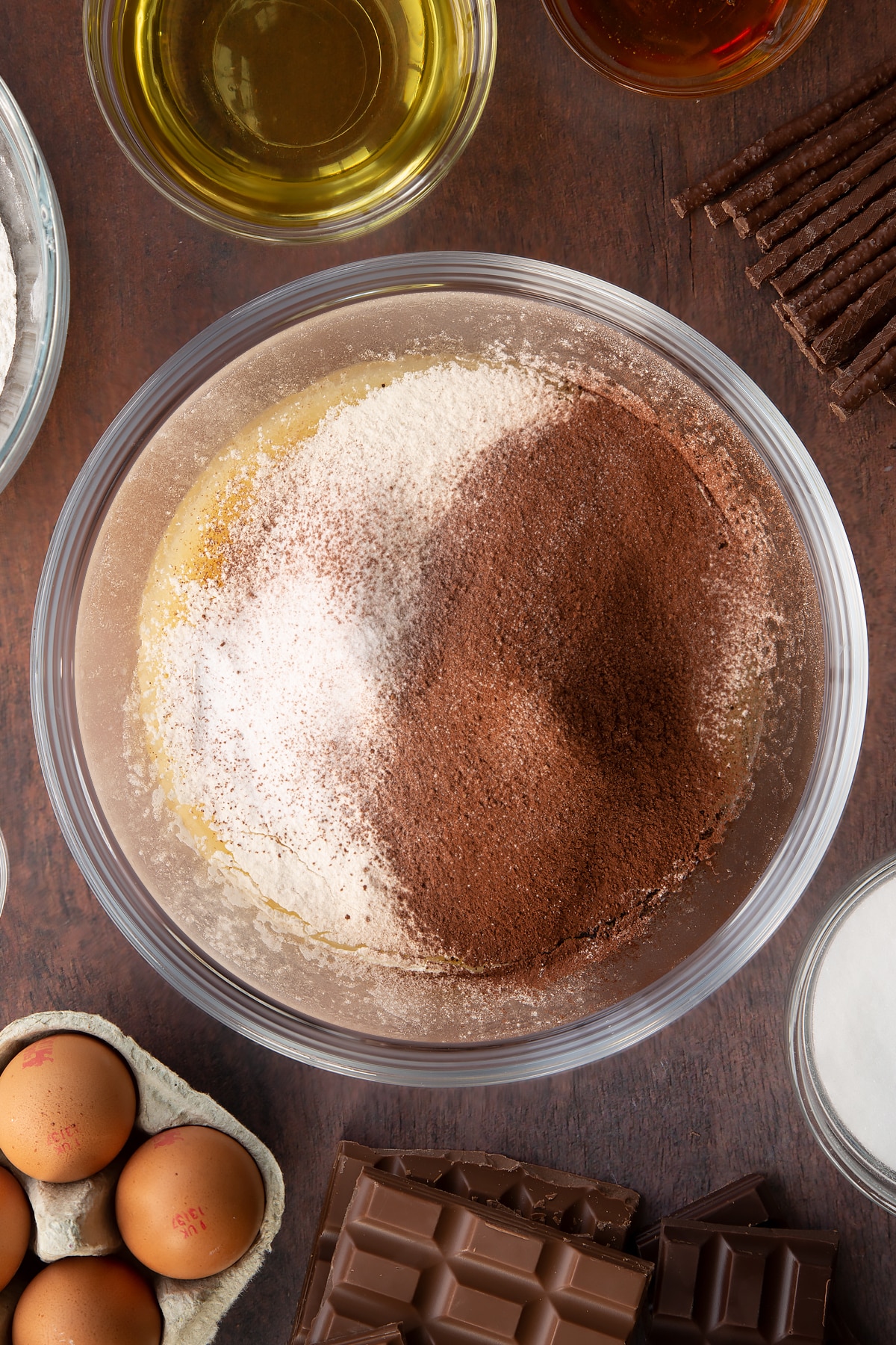 Sugar, beaten eggs, oil, milk and golden syrup, whisked together in a glass mixing bowl with flour, bicarb and cocoa on top. Ingredients to make Matchmaker cake surround the bowl.