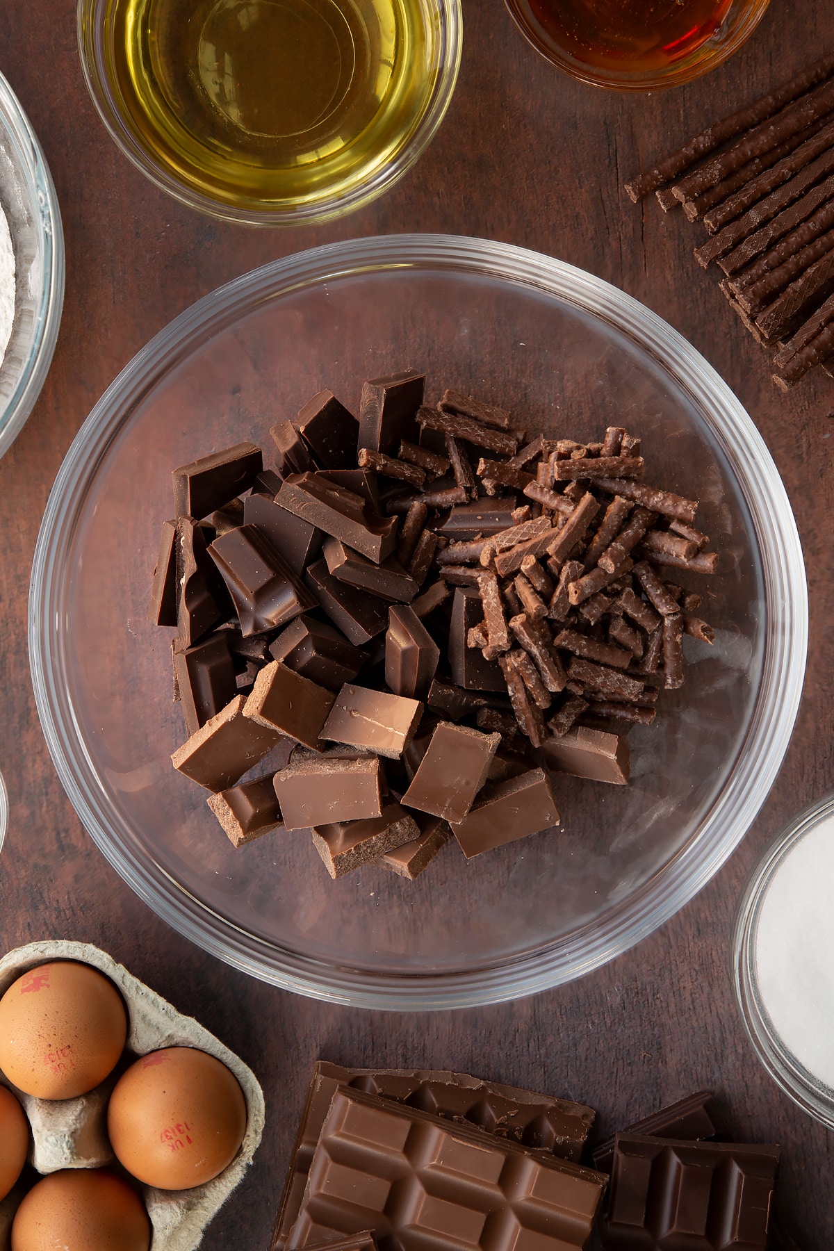 Dark chocolate, milk chocolate and Matchmakers in a glass mixing bowl. Ingredients to make Matchmaker cake surround the bowl.