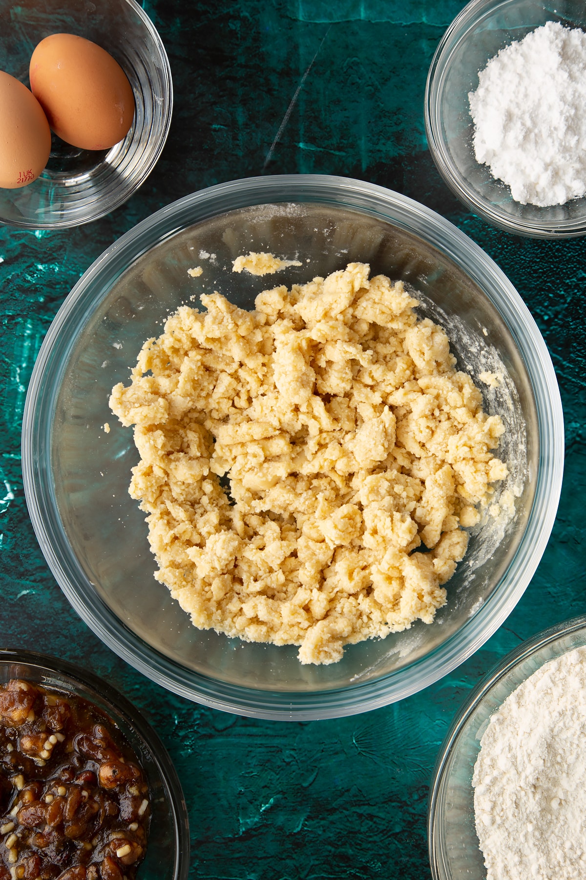 Roughly mixed pastry in a mixing bowl. Ingredients to make mini mince pies surround the bowl.