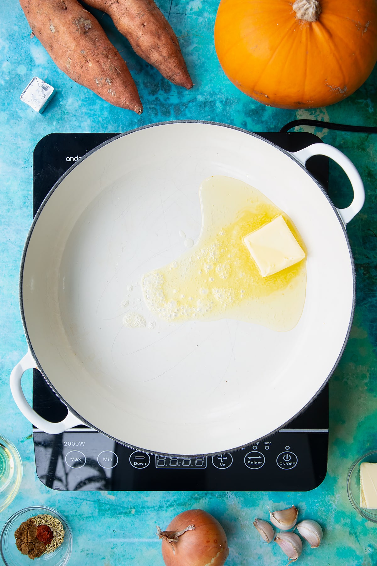 Butter and oil melting in a large, shallow pan, from above. Ingredients to make pumpkin and sweet potato soup surround the pan.