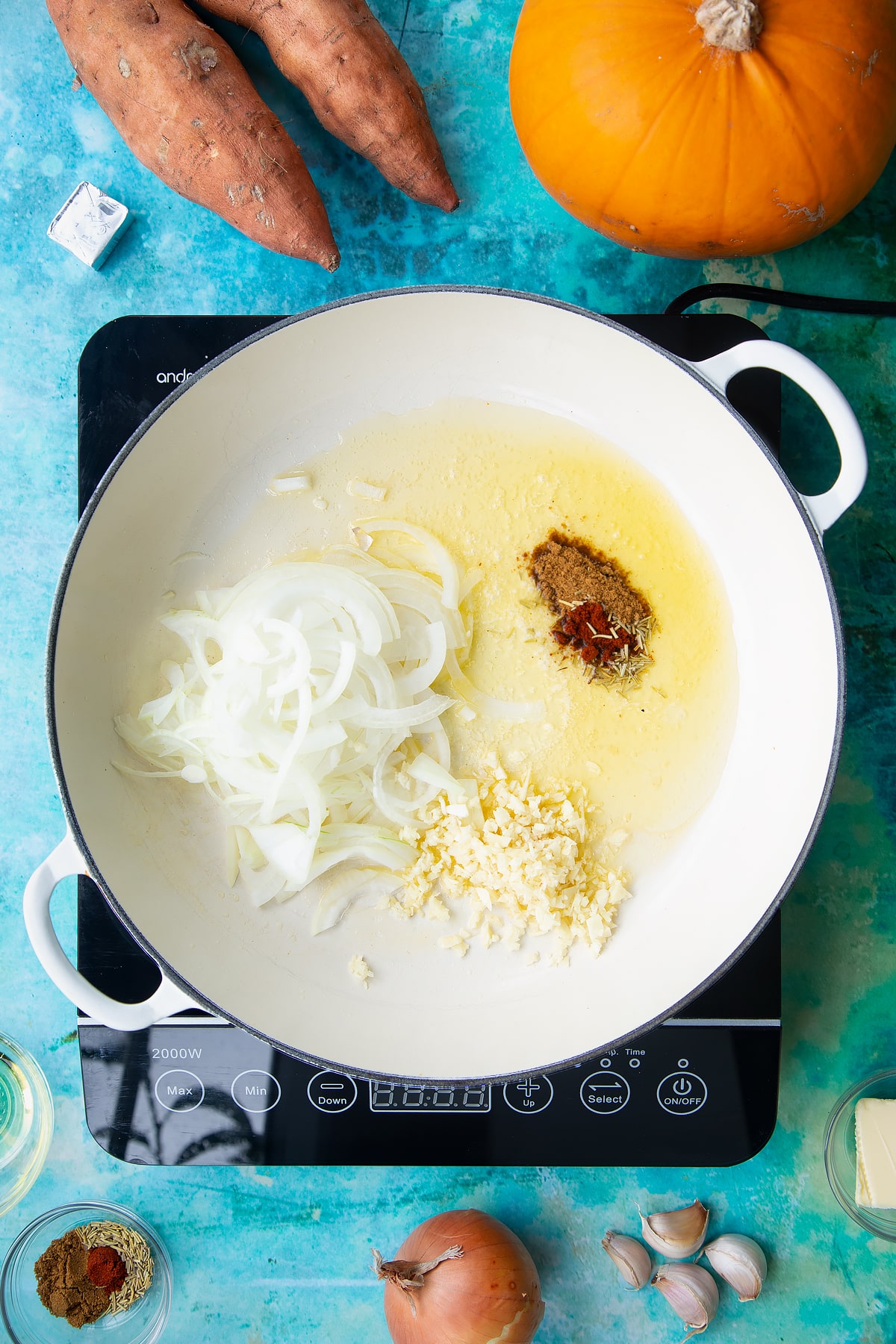 Onion, garlic, herbs and spices in a large, shallow pan, from above. Ingredients to make pumpkin and sweet potato soup surround the pan.