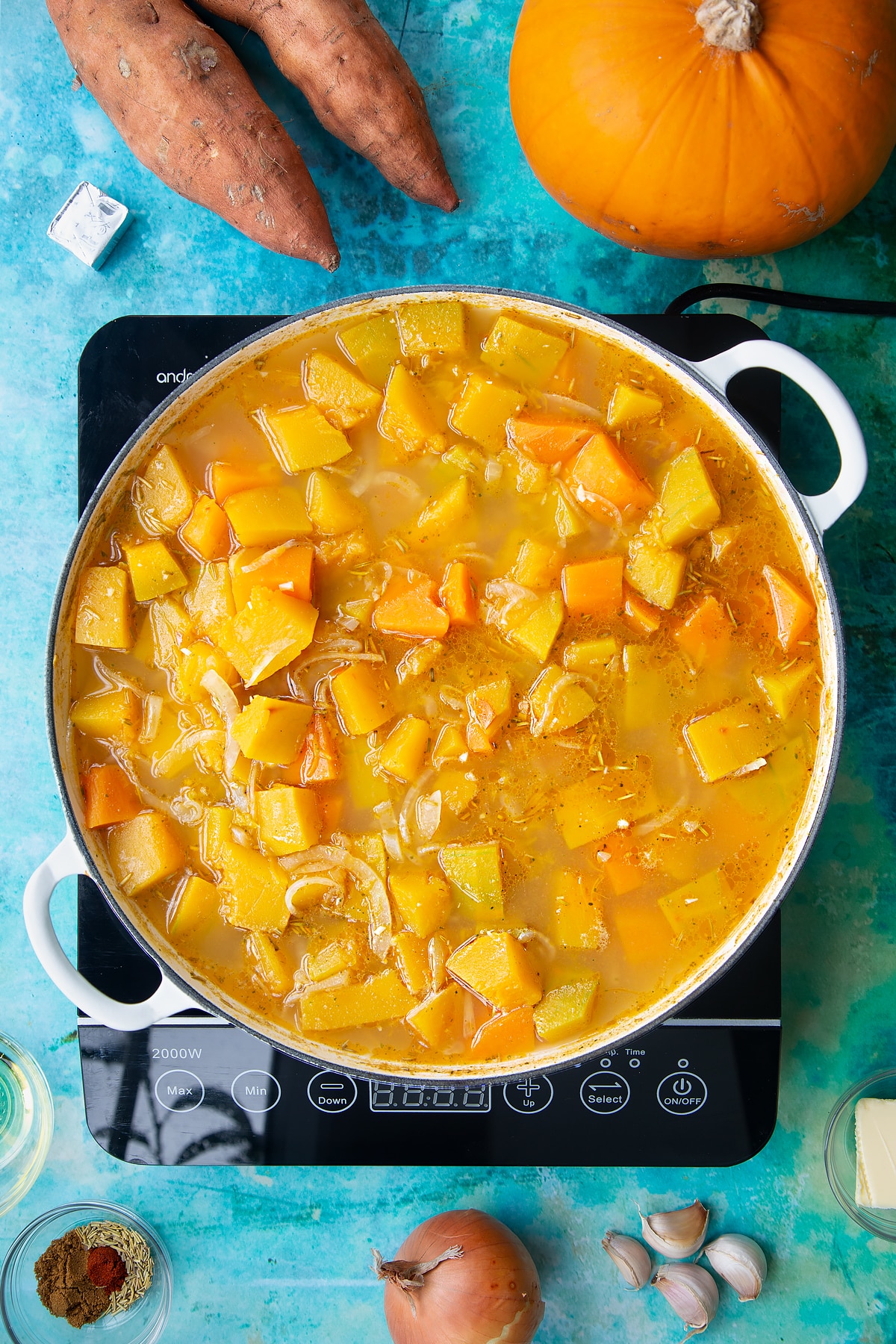 Pumpkin, sweet potato, onion and garlic cooked in stock in a large, shallow pan, from above. Ingredients to make pumpkin and sweet potato soup surround the pan.
