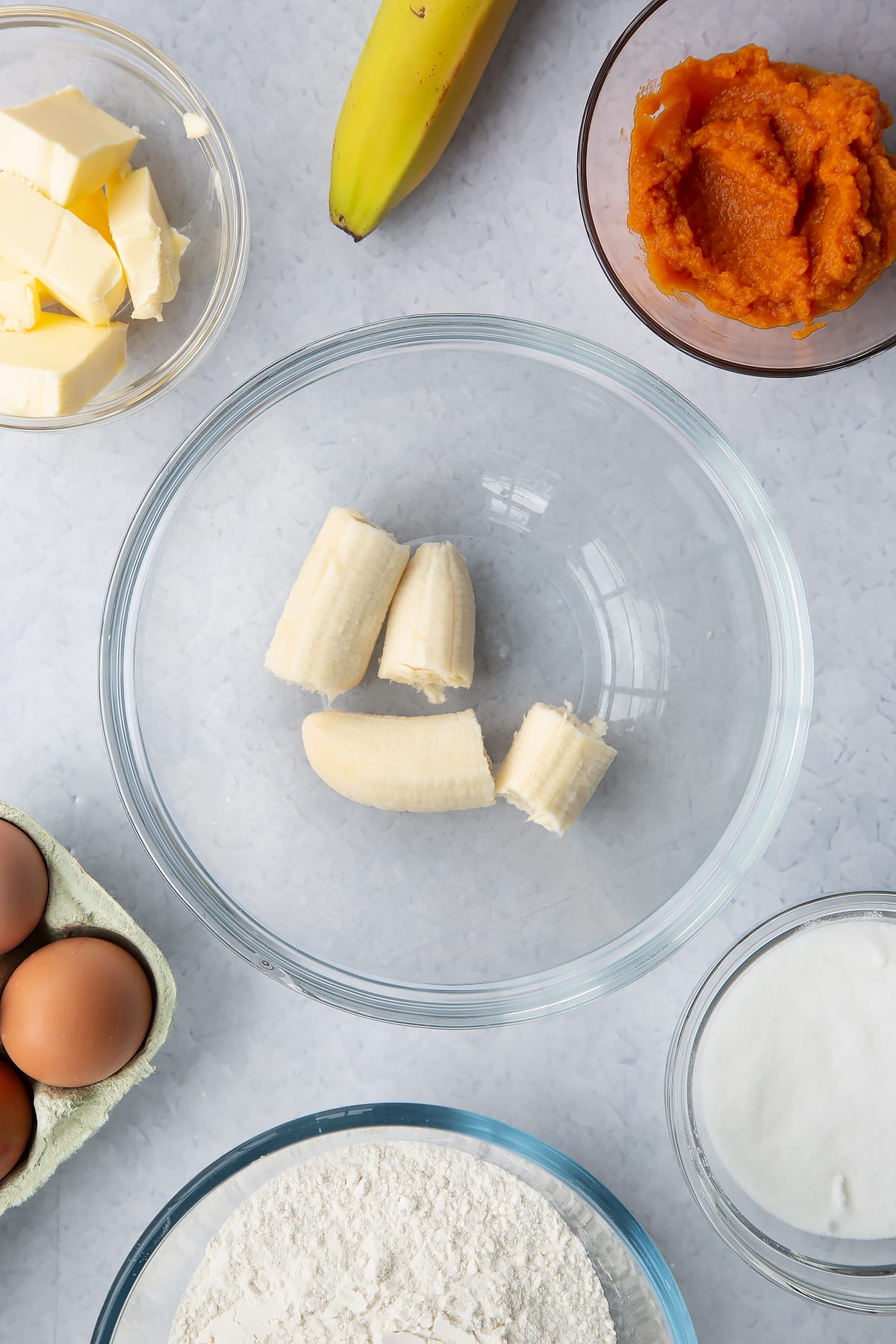 Banana in a glass mixing bowl. Ingredients to make pumpkin banana bread surround the bowl.