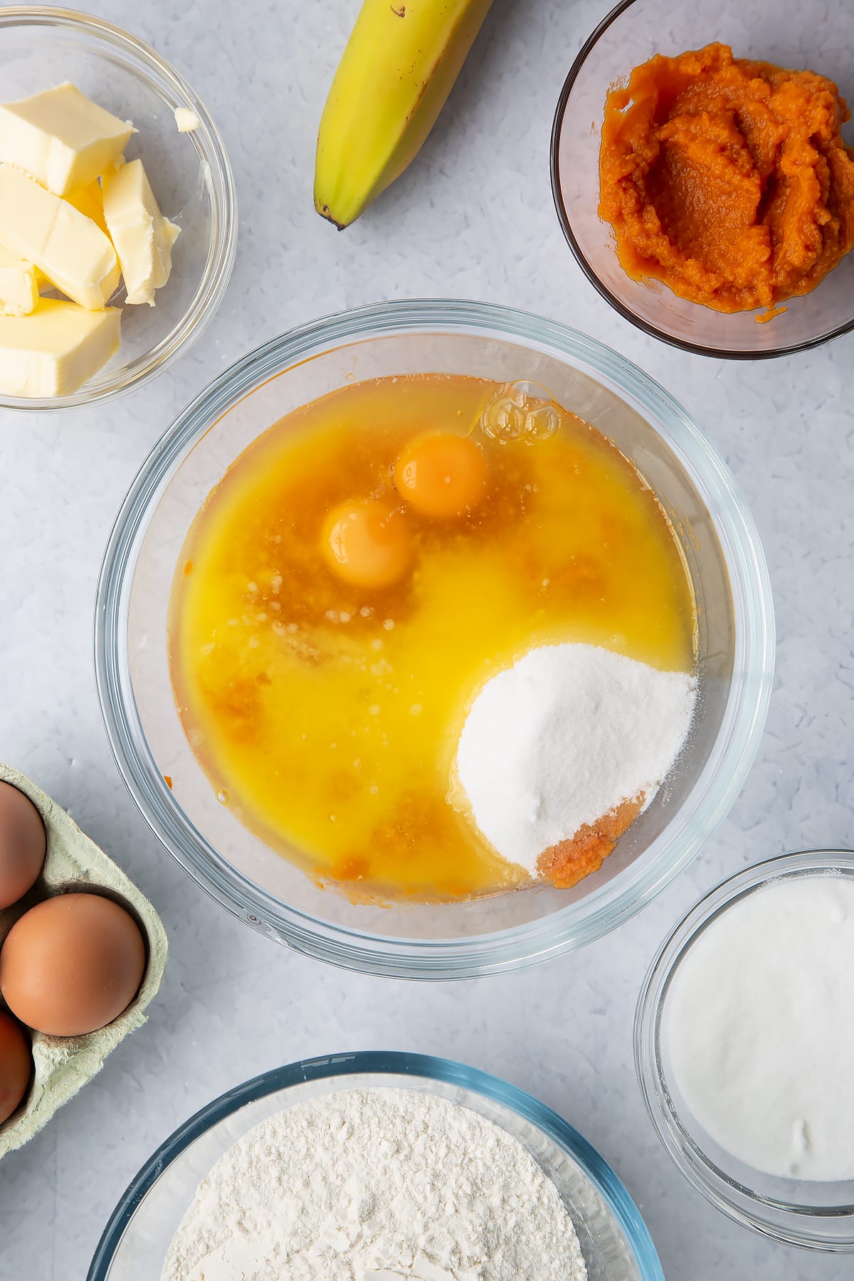 Mashed banana mixed with pumpkin puree in a glass mixing bowl with eggs, sugar and melted butter on top. Ingredients to make pumpkin banana bread surround the bowl.