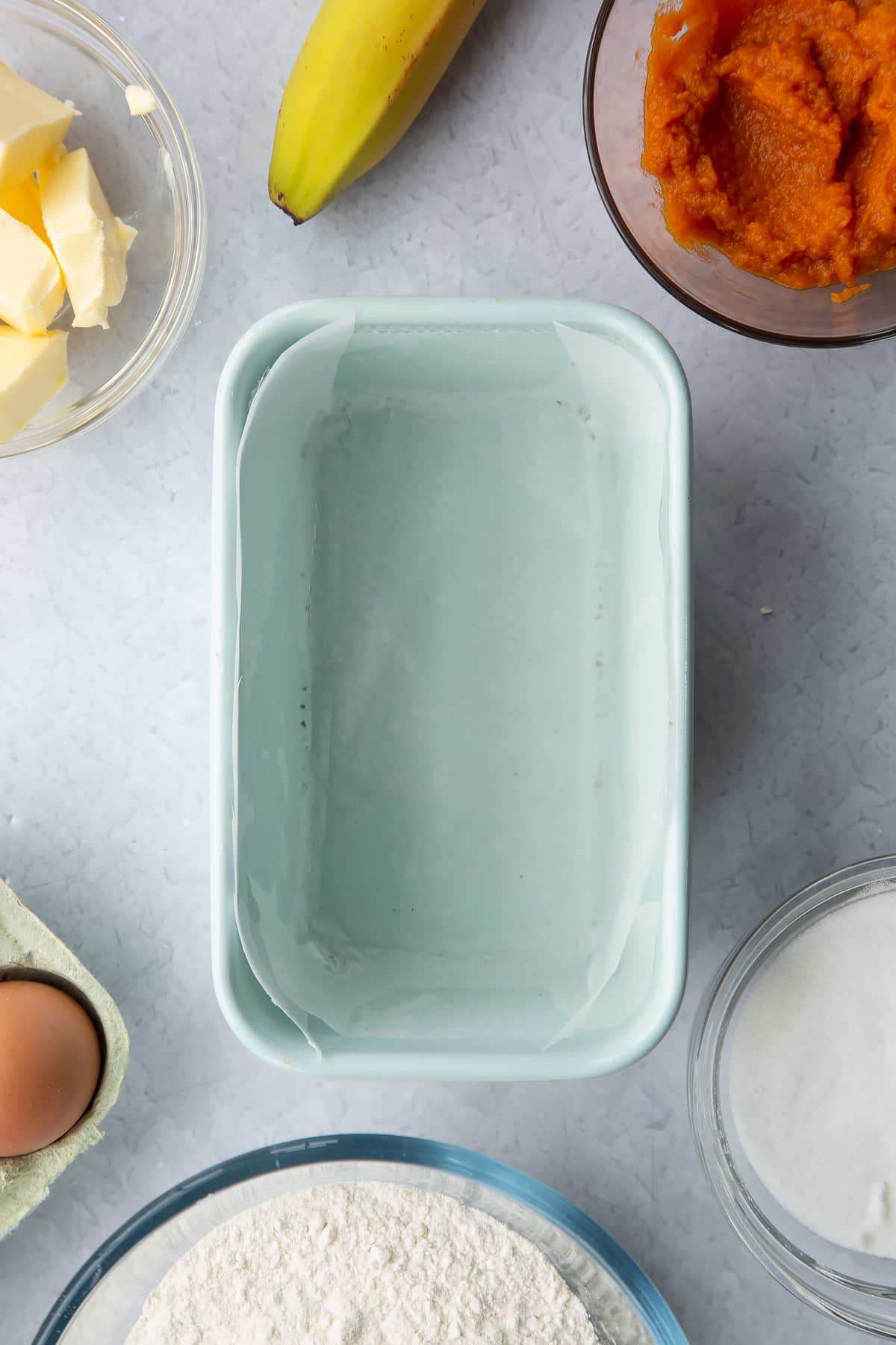 Loaf tin lined with baking paper, surrounded by ingredients to make pumpkin banana bread.