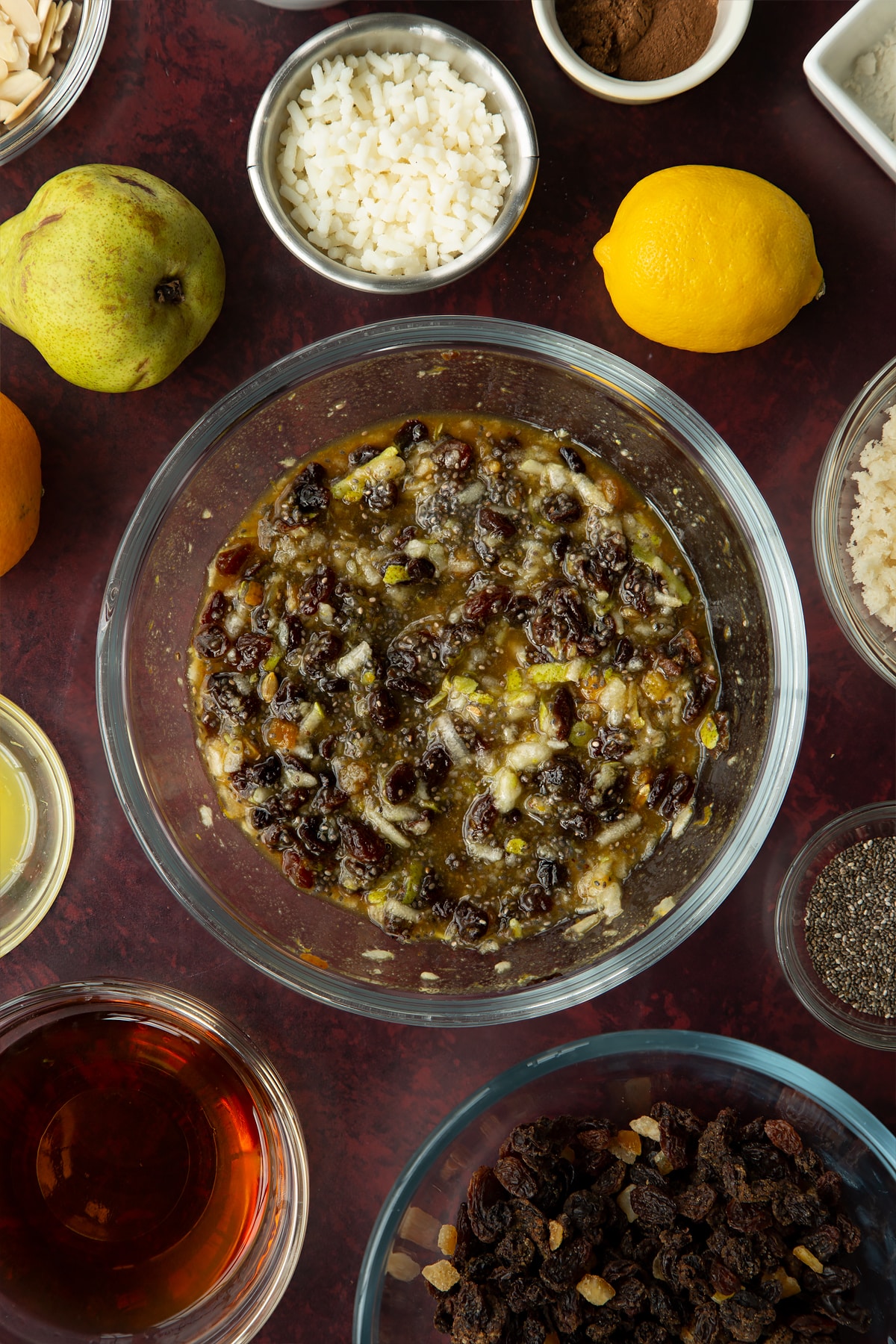 Plump mixed fruit, pear, soaked chia seeds, black treacle and sugar mixed together in a glass bowl. Ingredients to make vegan Christmas pudding surround the bowl.