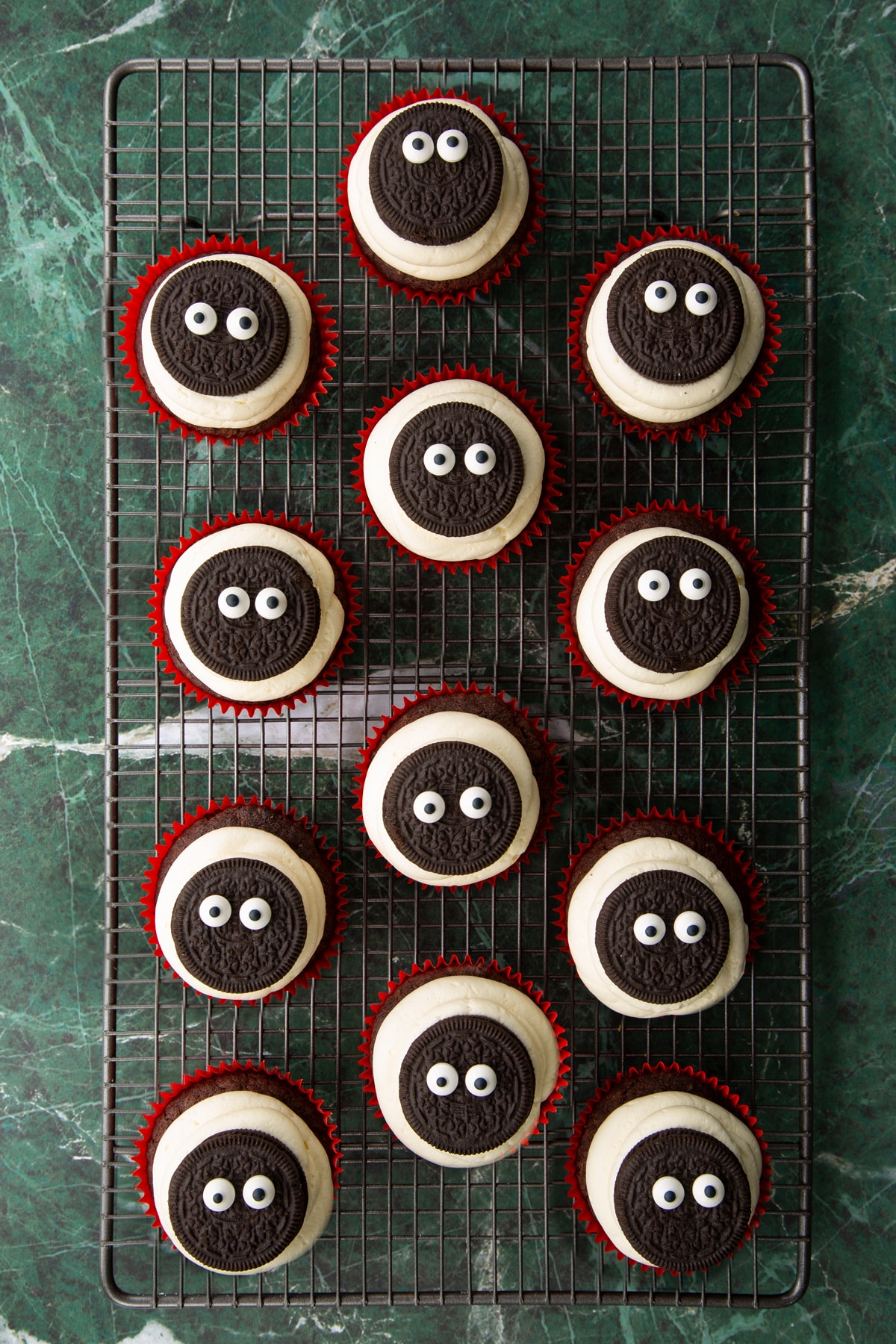 Vegan chocolate cupcakes on a wire cooling rack. They have vanilla frosting piped on top, then an Oreo with candy eyes.