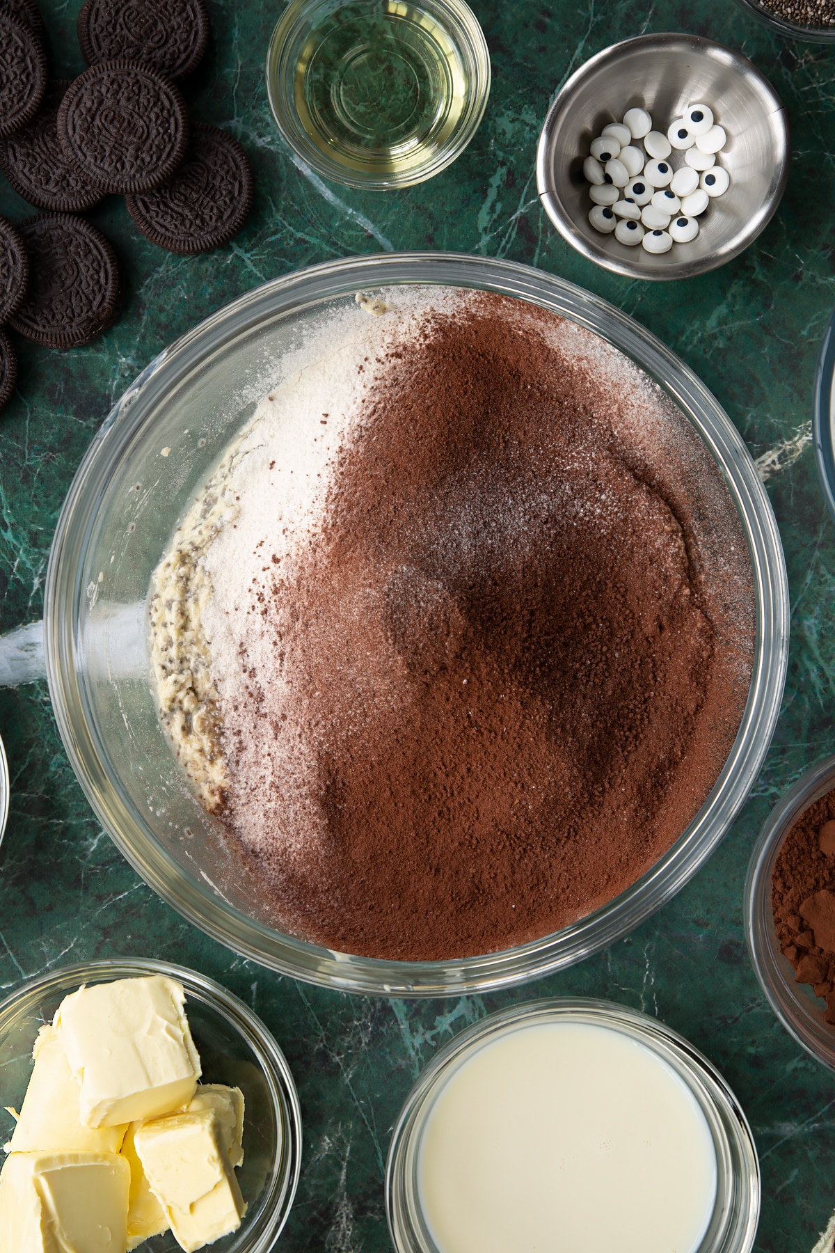 Vegan butter, caster sugar, chia "egg" and soya milk beaten in a glass mixing bowl, with cocoa, flour and baking powder on top. Ingredients to make vegan Halloween cupcakes surround the bowl.