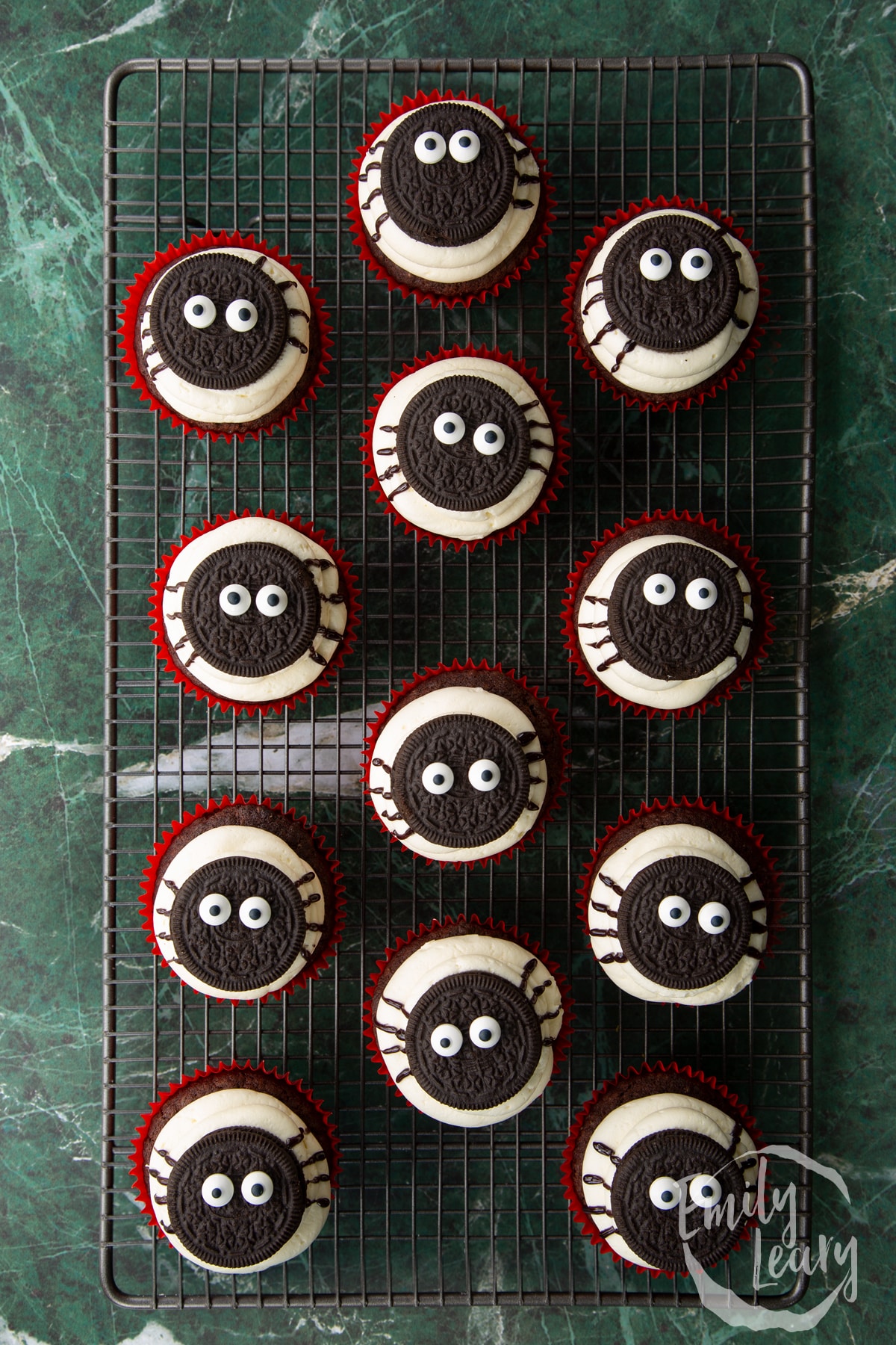 Vegan chocolate cupcakes on a wire cooling rack. They have vanilla frosting piped on top, then an Oreo with candy eyes. Spider legs are piped on with a black icing pen.