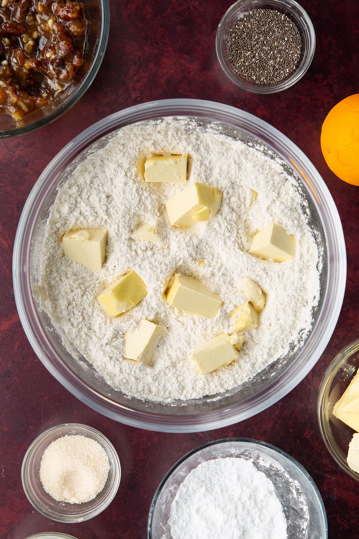 Flour and icing sugar mixed together in a bowl with cubes of vegan butter. Ingredients to make a vegan mince pie recipe surround the bowl.