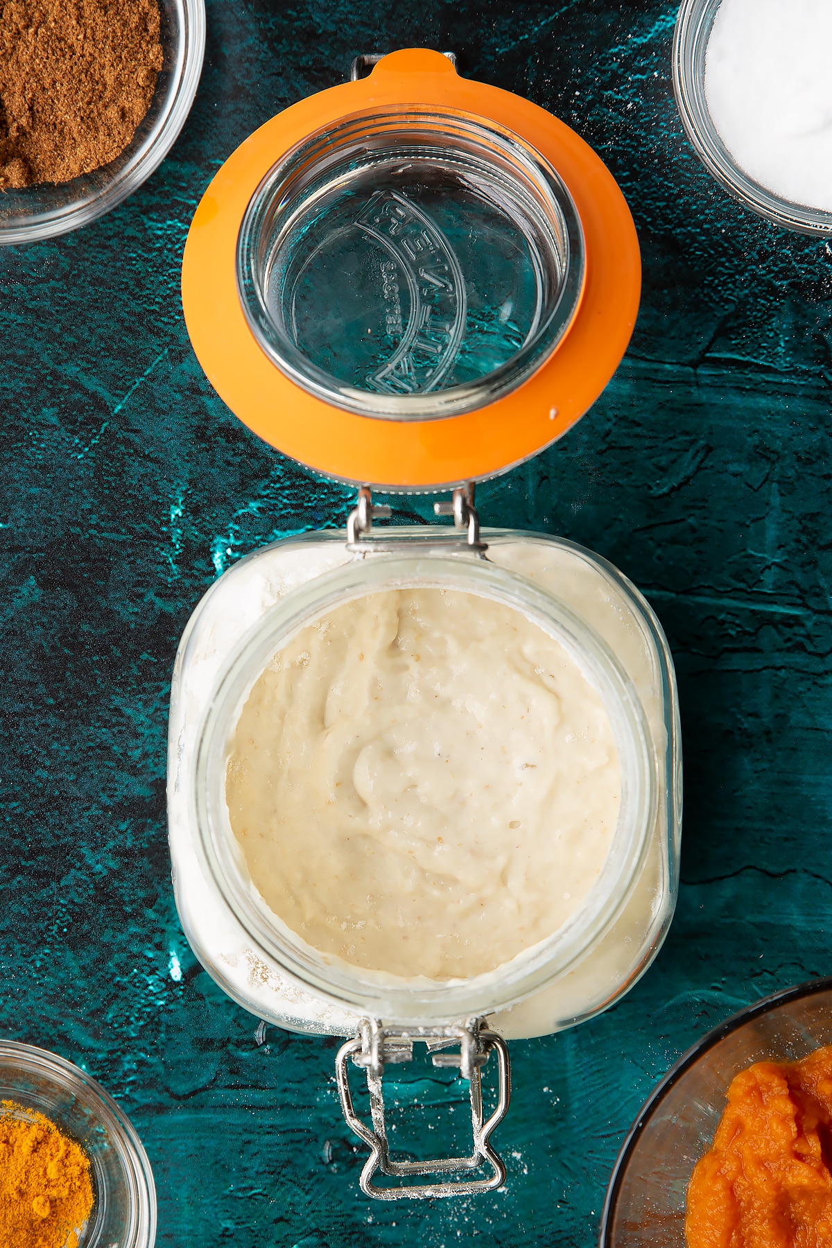 Sourdough starter in a jar with flour and water just stirred in. Ingredients to make pumpkin sourdough bread surround the jar.