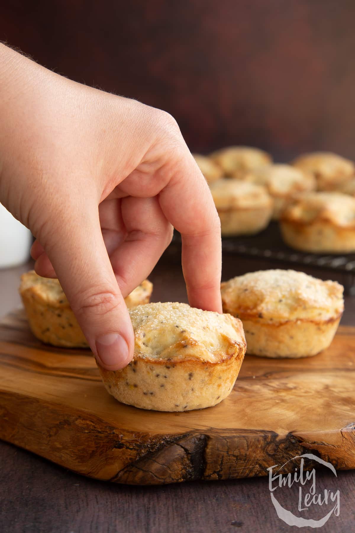 Vegan mince pies on a olive board with more on a wire rack in the background. A hand reaches for one.