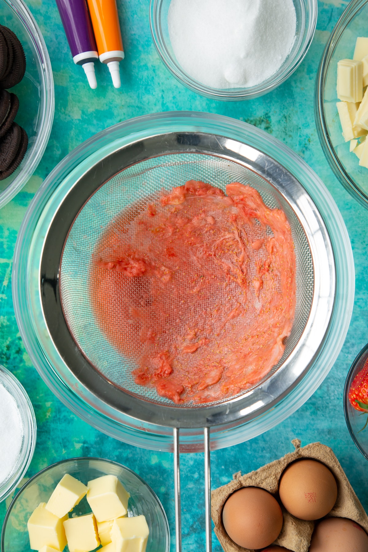 Strawberry pulp in a sieve over a bowl. Ingredients to make Halloween cheesecake surround the bowl.