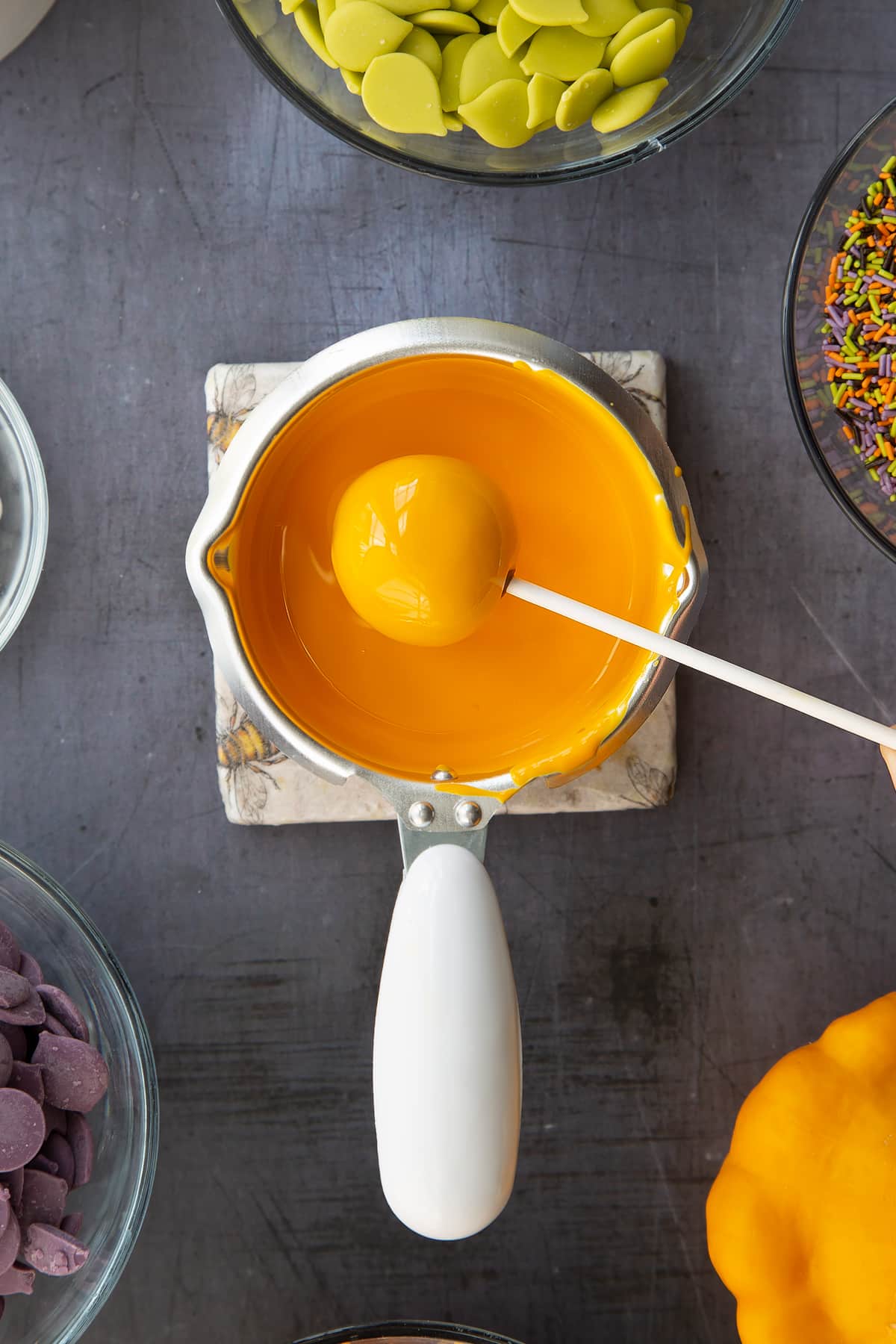 Orange candy melts in a little pan, a cake pop is being dipped into it. Ingredients to make Halloween cake pops surround the pan.
