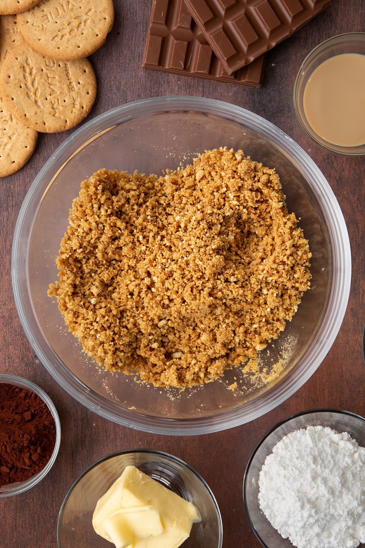 Digestive biscuit crumbs and melted butter mixed together in a glass bowl. Ingredients to make Baileys cheesecake surround the bowl.