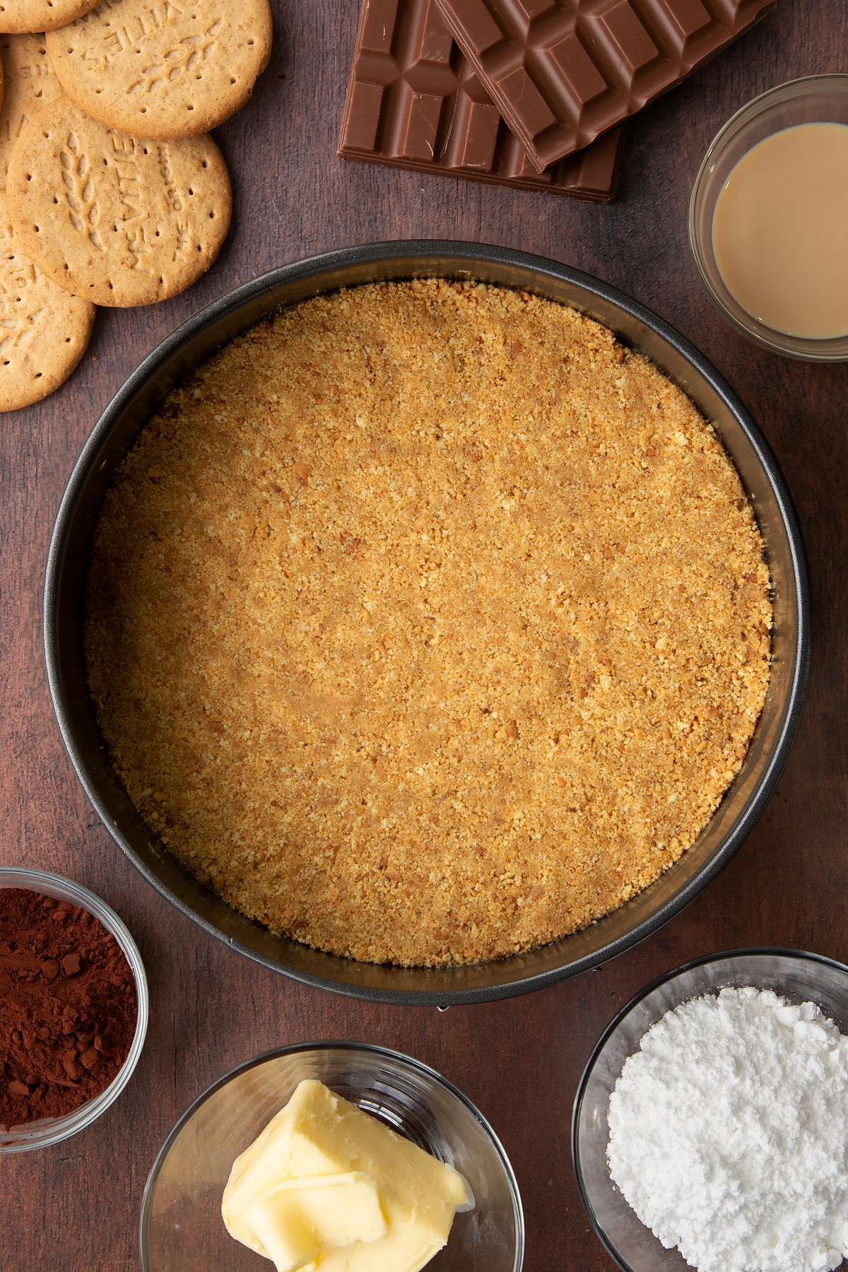 Digestive biscuit crumbs and melted butter mixed together and pressed into the base of a cake tin. Ingredients to make Baileys cheesecake surround the tin.