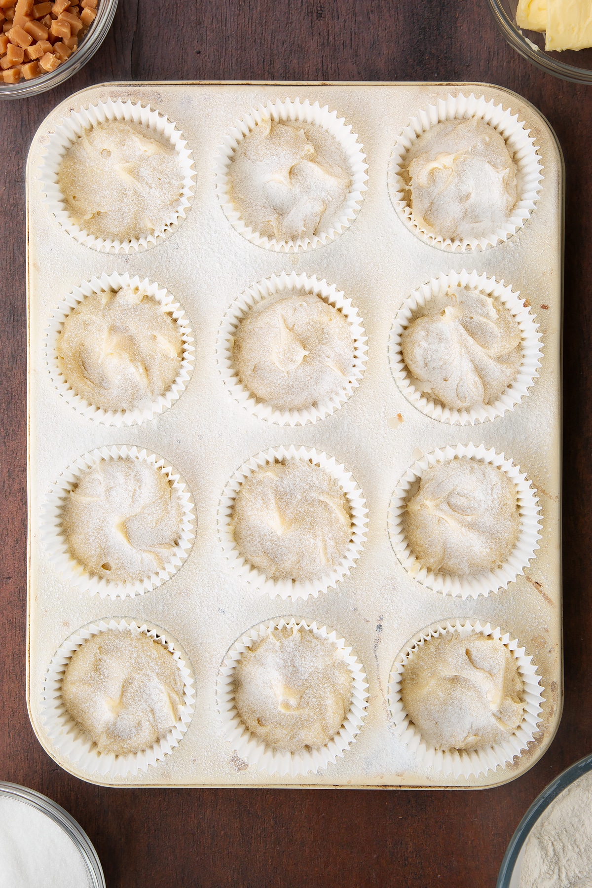 12 bun cases in a metal bun tray filled with vanilla cake batter topped with a light dusting on flour