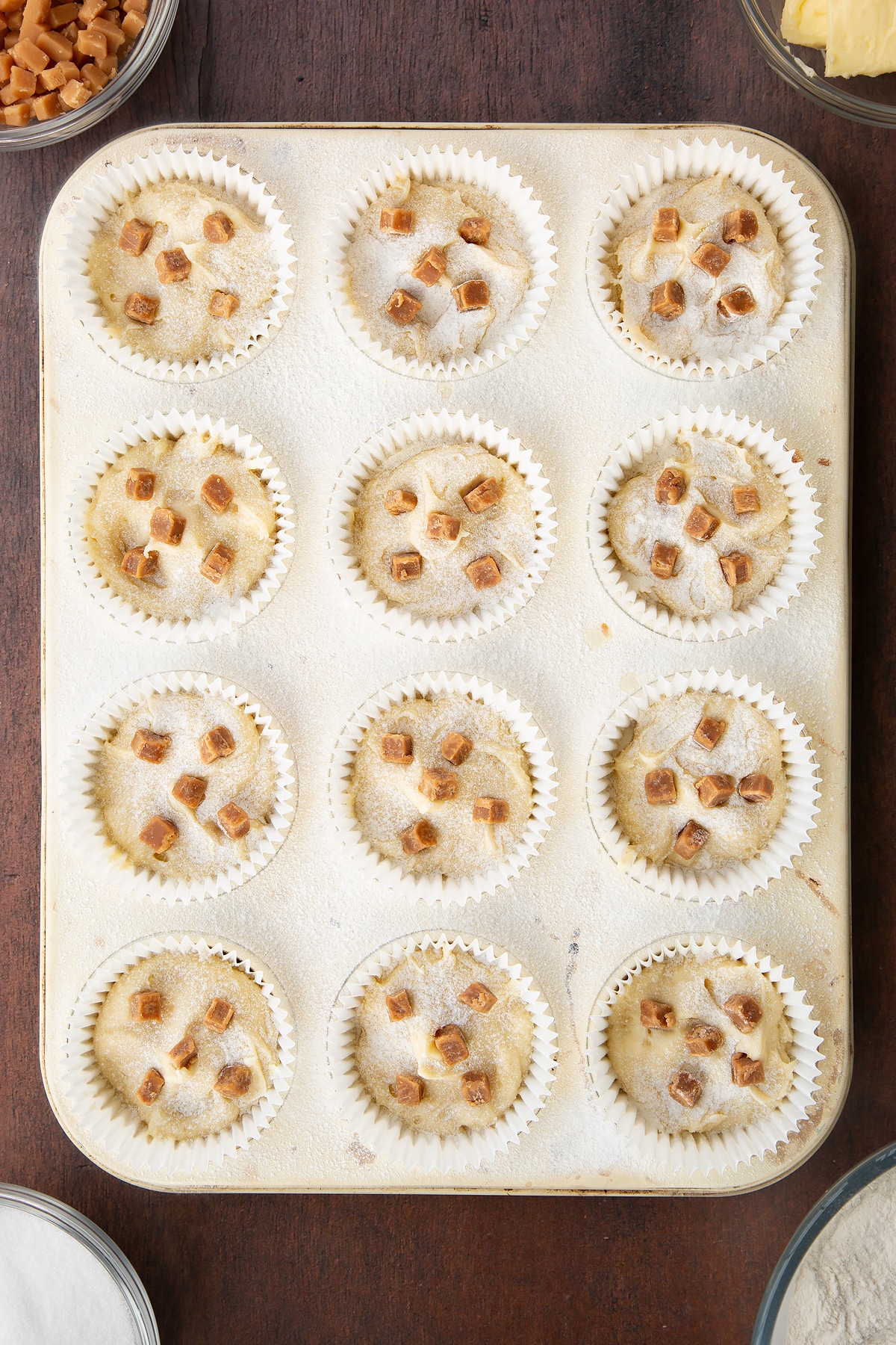 12 bun cases in a metal bun tray filled with vanilla cake batter topped with a light dusting on flour and fudge chips.