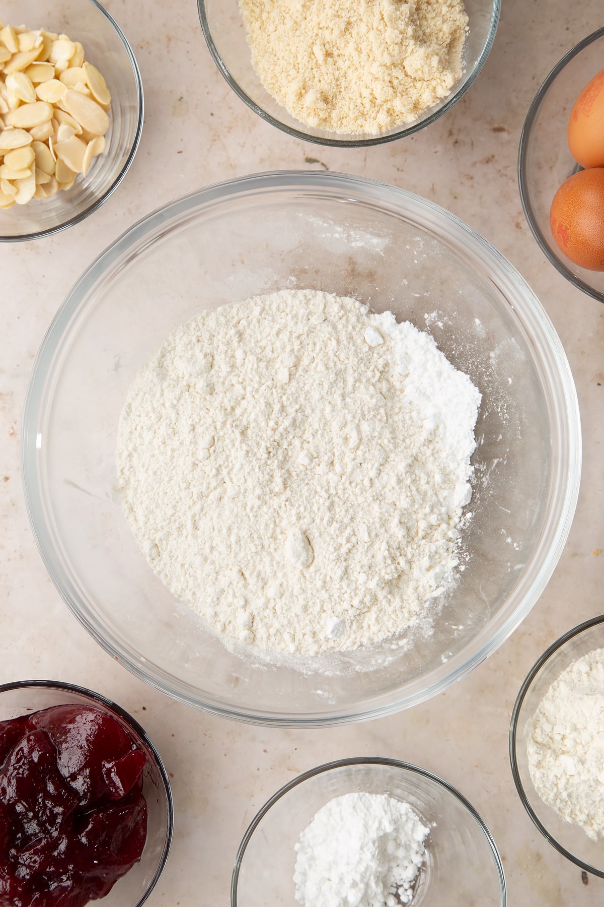 Flour and icing sugar in a bowl. Ingredients to make a Bakewell tray bake surround the bowl.