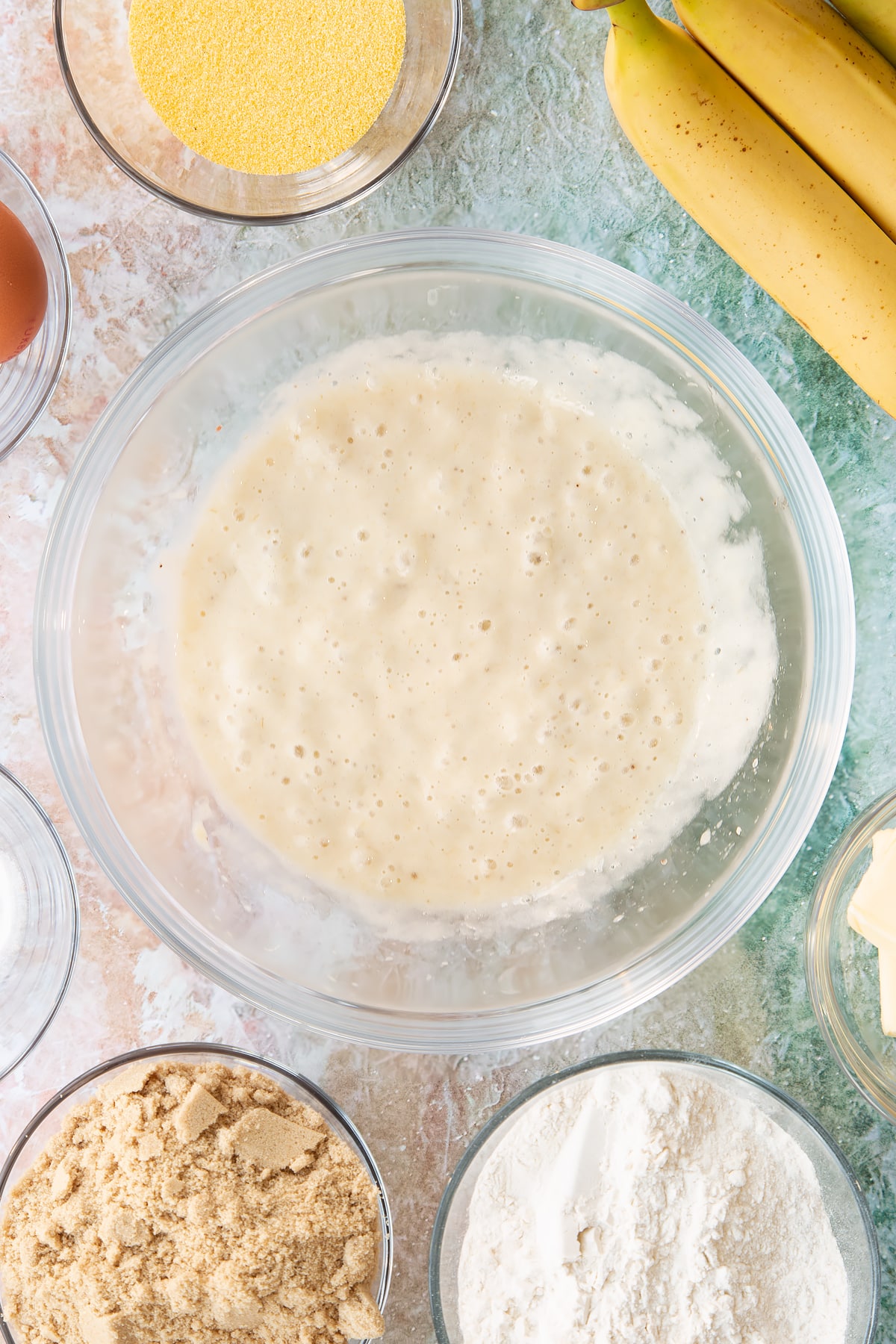 Mashed bananas in a bowl. Ingredients to make banana cornmeal muffin surround the bowl.