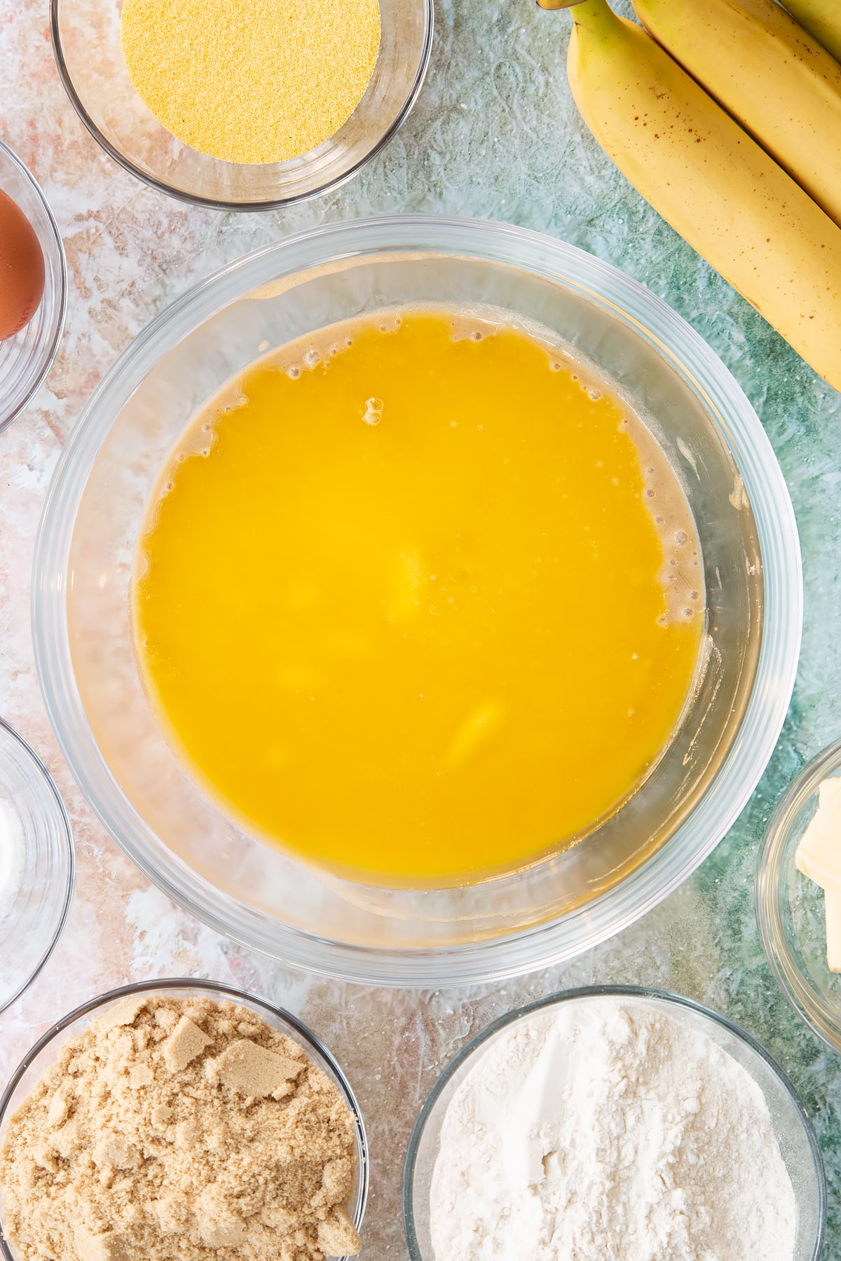 Mashed bananas and sugar mixed together in a bowl with melted butter on top. Ingredients to make banana cornmeal muffin surround the bowl.