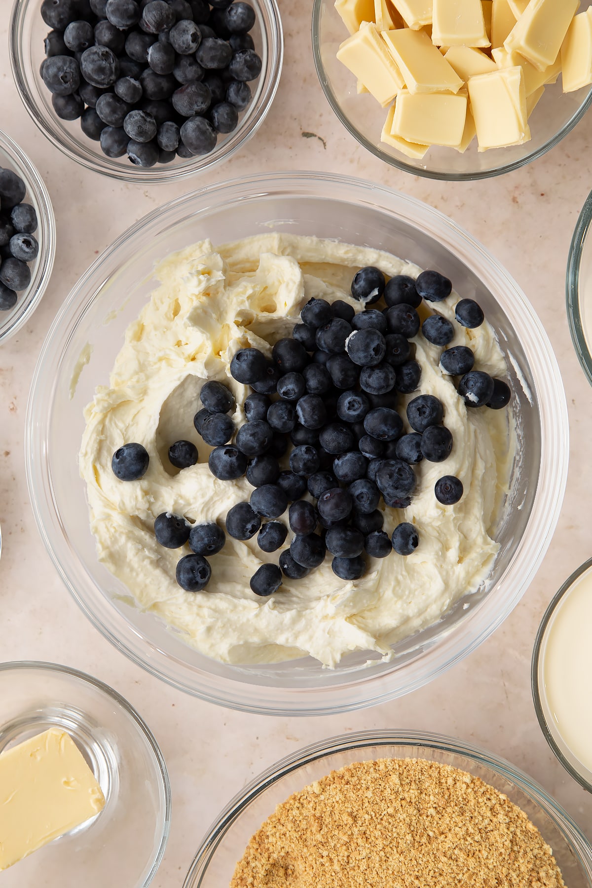 Cheesecake filling in a bowl with blueberries on top. Ingredients to make blueberry cheesecake pie surround the bowl.