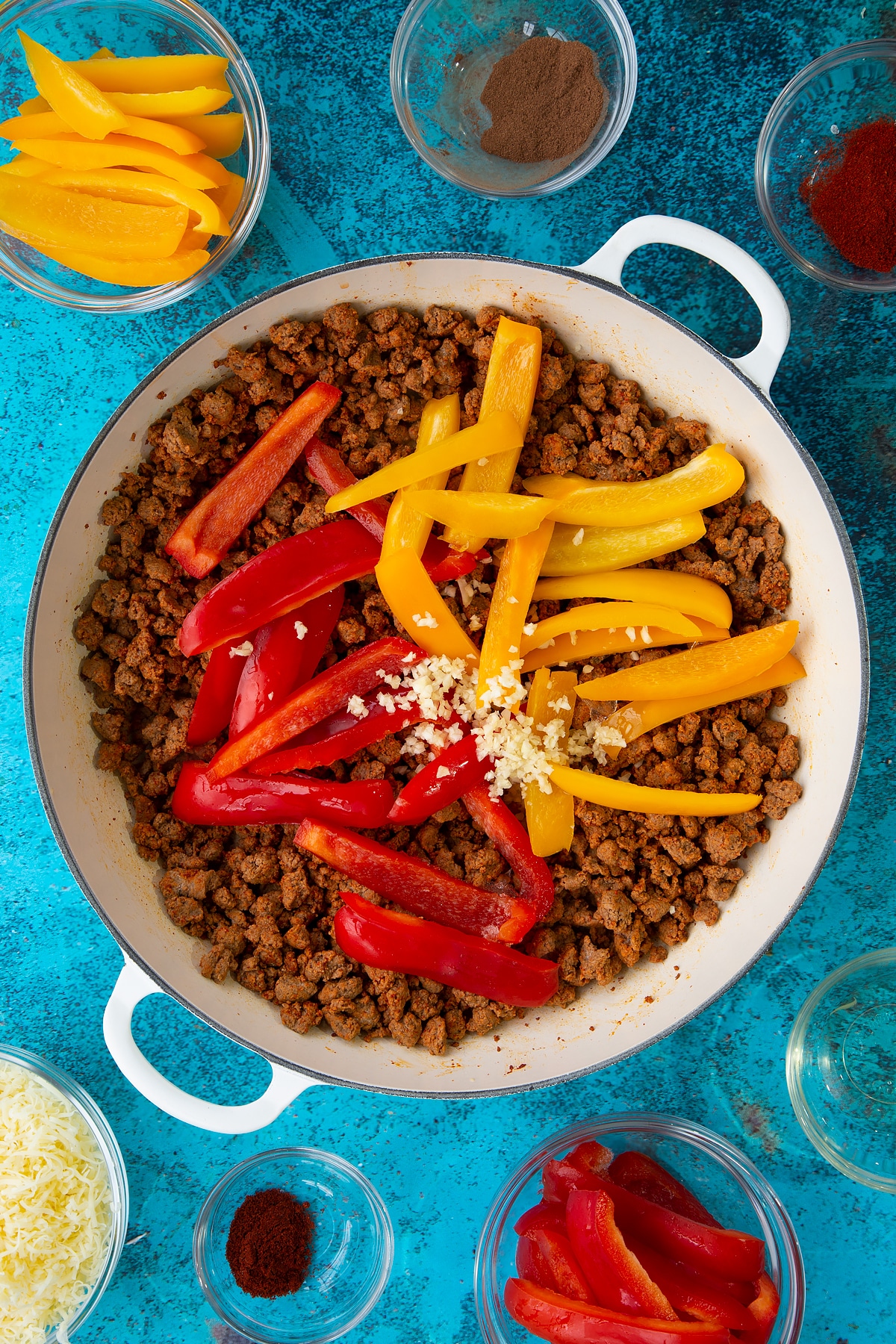 Quorn mince fried in spices in a large white pan with garlic and peppers on top. Ingredients to make Quorn mince enchiladas surround the pan.