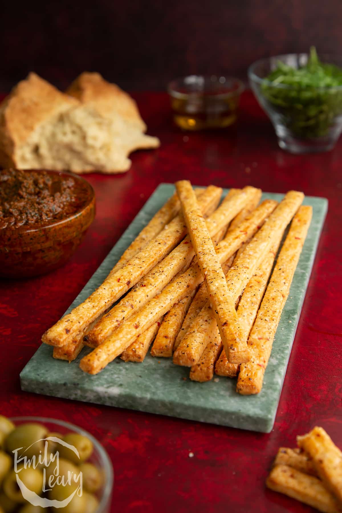 A batch of spicy cheese straws on a decorative board with garnish in the background.