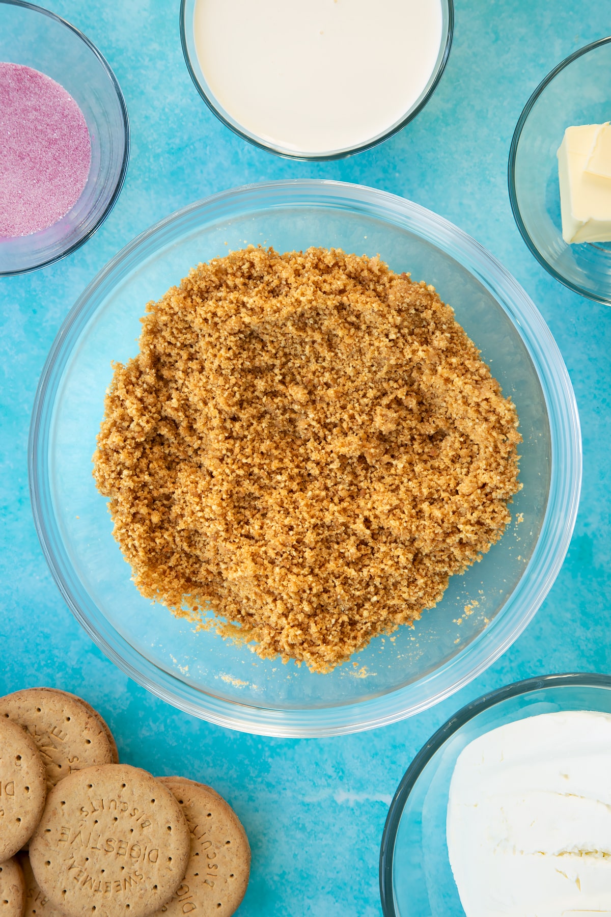 Crushed biscuits mixed with melted butter in a glass bowl. Ingredients to make jello cheesecake surround the bowl.