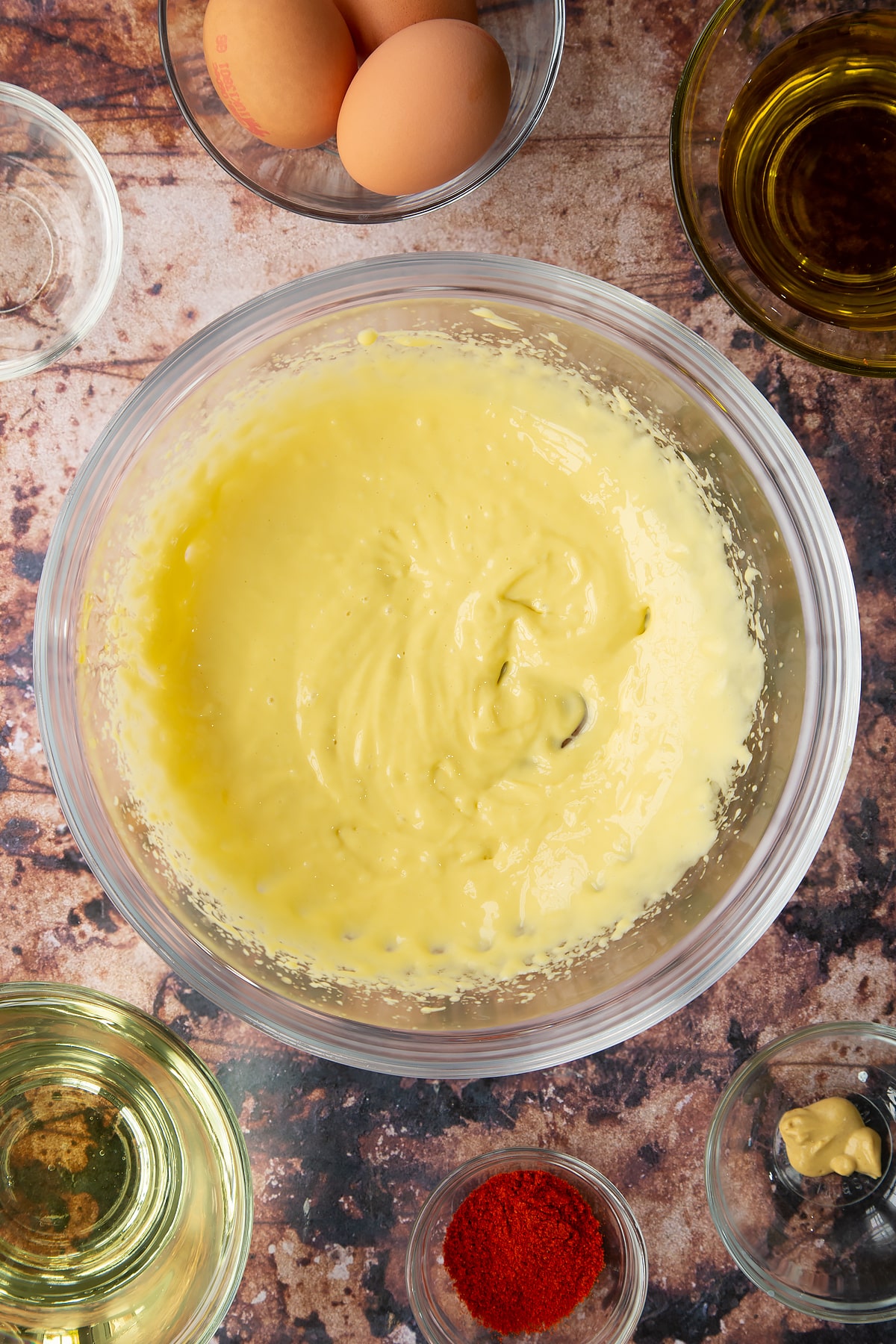 A small amount of thin mayo just beginning to come together in a bowl. Ingredients to make paprika mayo surround the bowl.