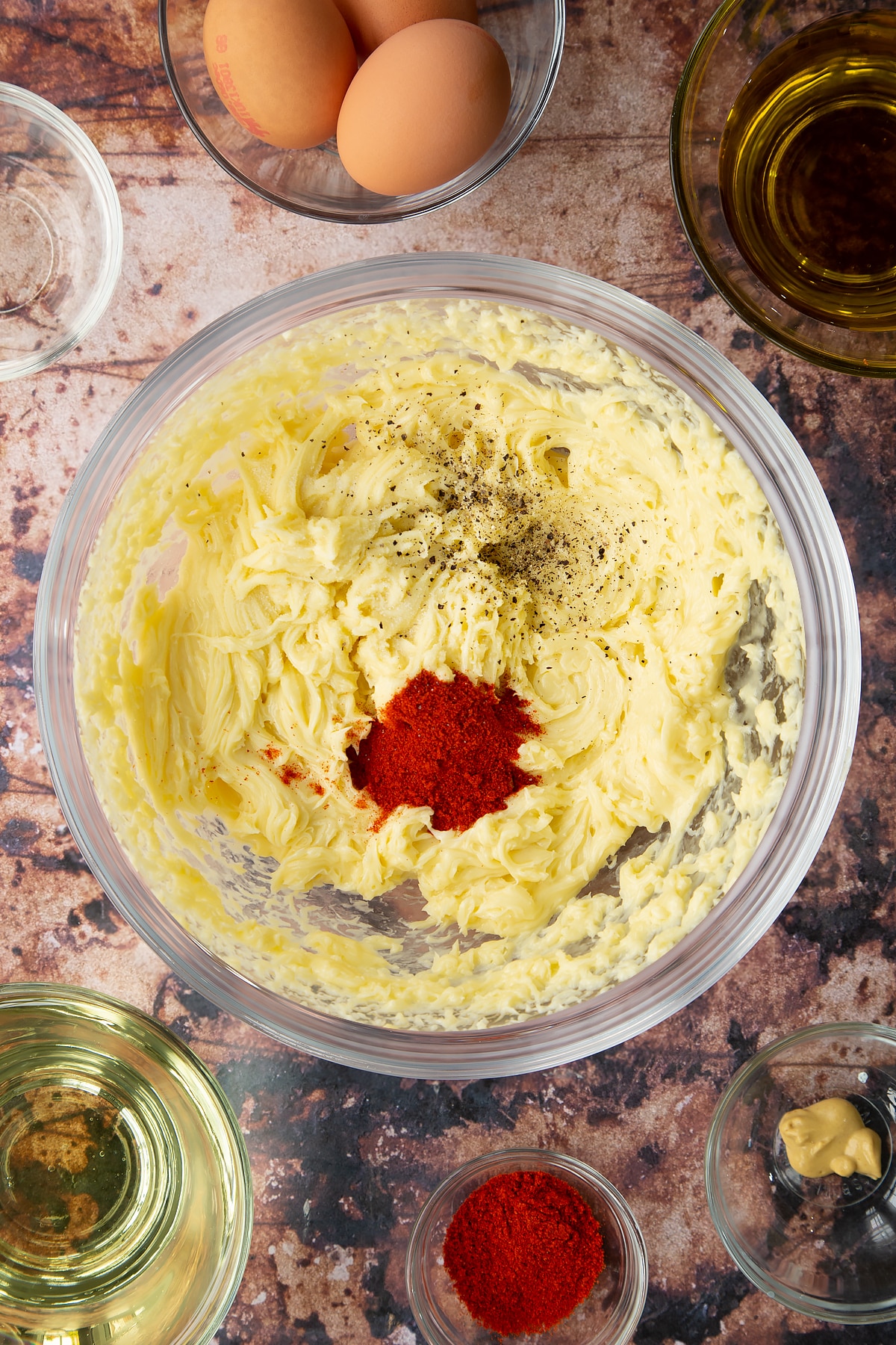 Thick olive oil mayo in a bowl with paprika and pepper on top. Ingredients to make paprika mayo surround the bowl.
