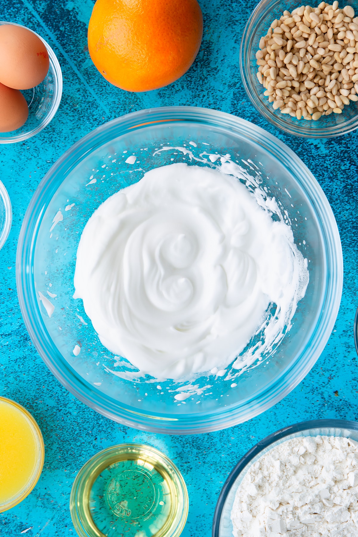 Stiffly beaten eggs whites in a bowl. Ingredients to make pine nut cake surround the bowl.