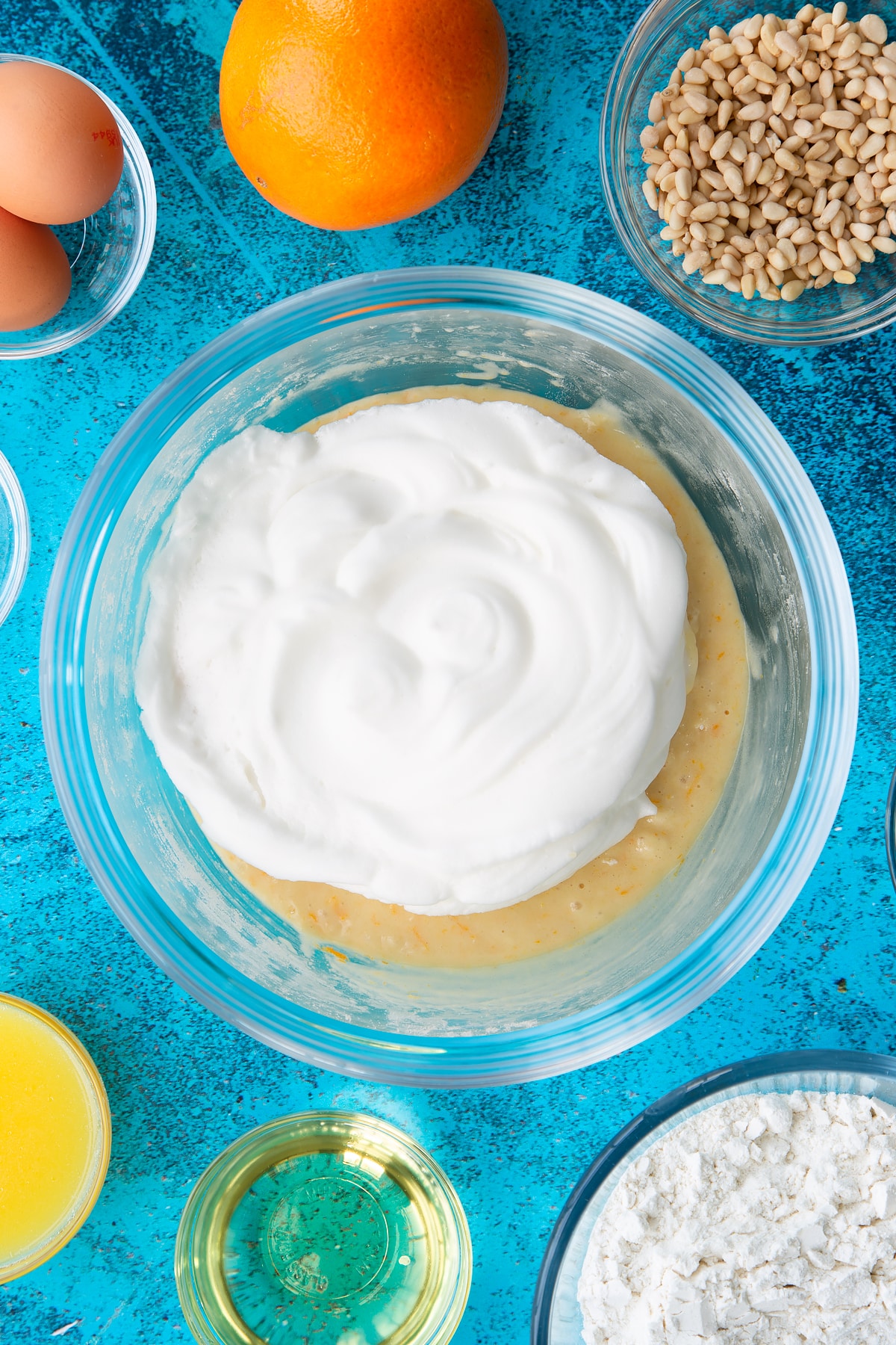 Sugar, egg yolks, oil, orange juice, zest, flour and baking powder whisked together in a bowl with beaten egg whites on top. Ingredients to make pine nut cake surround the bowl.